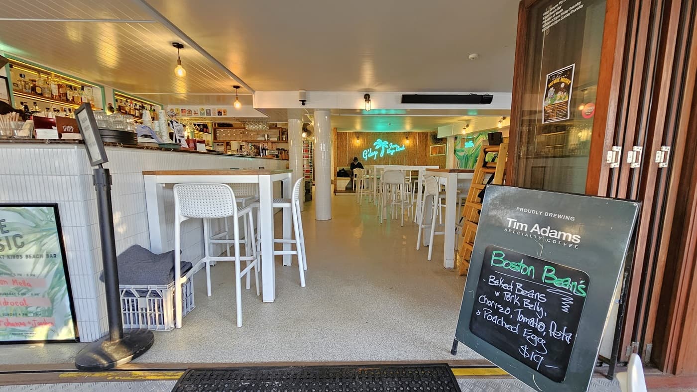 On the left, there is a white display freezer with an owl figurine on top and a glass display case containing food items. In the middle, there is an open entrance leading into a café with white tables and chairs, a counter with stools, and a neon sign on the back wall. On the right, there are more white tables and chairs, a mural of a parrot on the wall, and a wooden partition. The floor is concrete, and the ceiling has exposed wooden beams and white panels.