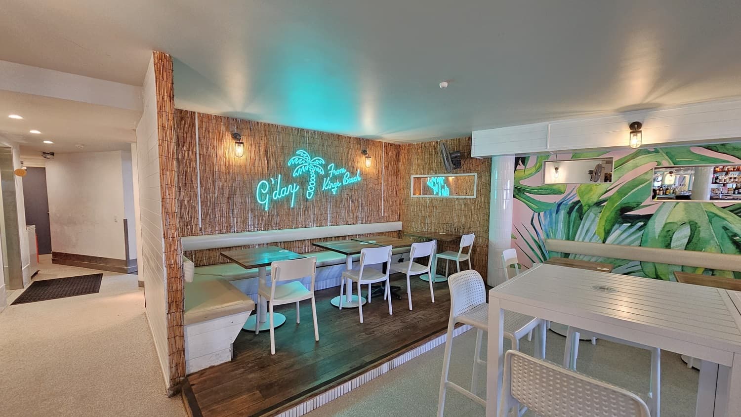On the left, there is a bamboo wall with a neon sign reading "G'day From Kings Beach" above a seating area with brown tables and beige cushioned benches. There is a wooden platform with additional seating and a small mirror on the wall. On the right, there is a mural of green leaves on the wall, a bar counter with stools, and shelves with bottles. The floor is light-coloured  and the ceiling is white with recessed lighting.A corridor leading to an area with white walls is visible on the left.