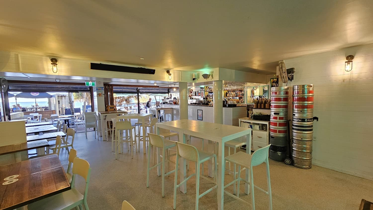 On the left, there are white tables and chairs near a wooden wall with a window. In the middle, there are high white tables with bar stools, and a bar counter with various bottles and glasses. In the background, there is an open area with more tables and chairs, and a view of the beach. On the right, there are stacked beer kegs and a white brick wall. The floor is light-coloured concrete, and the ceiling is white with recessed lighting and wall-mounted lamps.