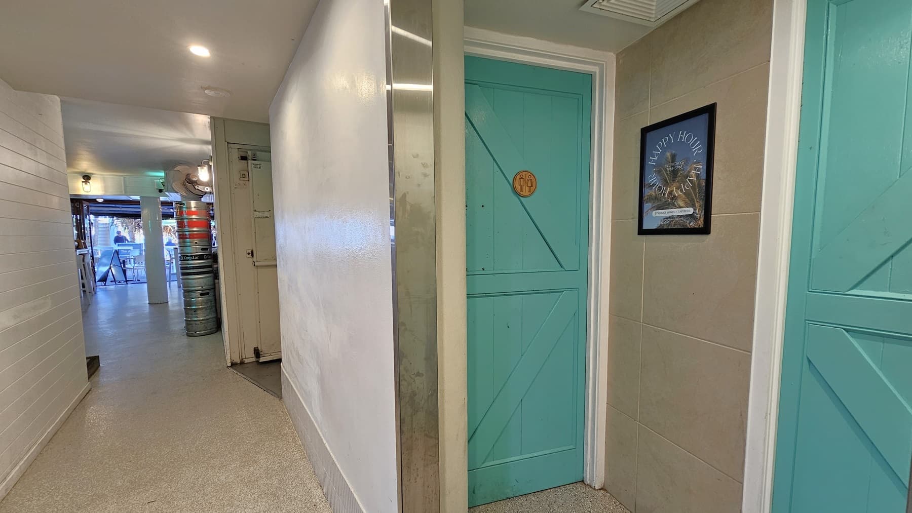 On the left, there is a hallway with white walls and a light-coloured floor, leading to an open area with stacked beer kegs and a view of tables and chairs. In the middle, there is a white wall with a narrow vertical mirror. On the right, there are two turquoise doors with wooden panels, one with a "WC" sign and the other with a framed picture of a beach scene. The floor is light-coloured concrete, and the ceiling is white with recessed lighting.