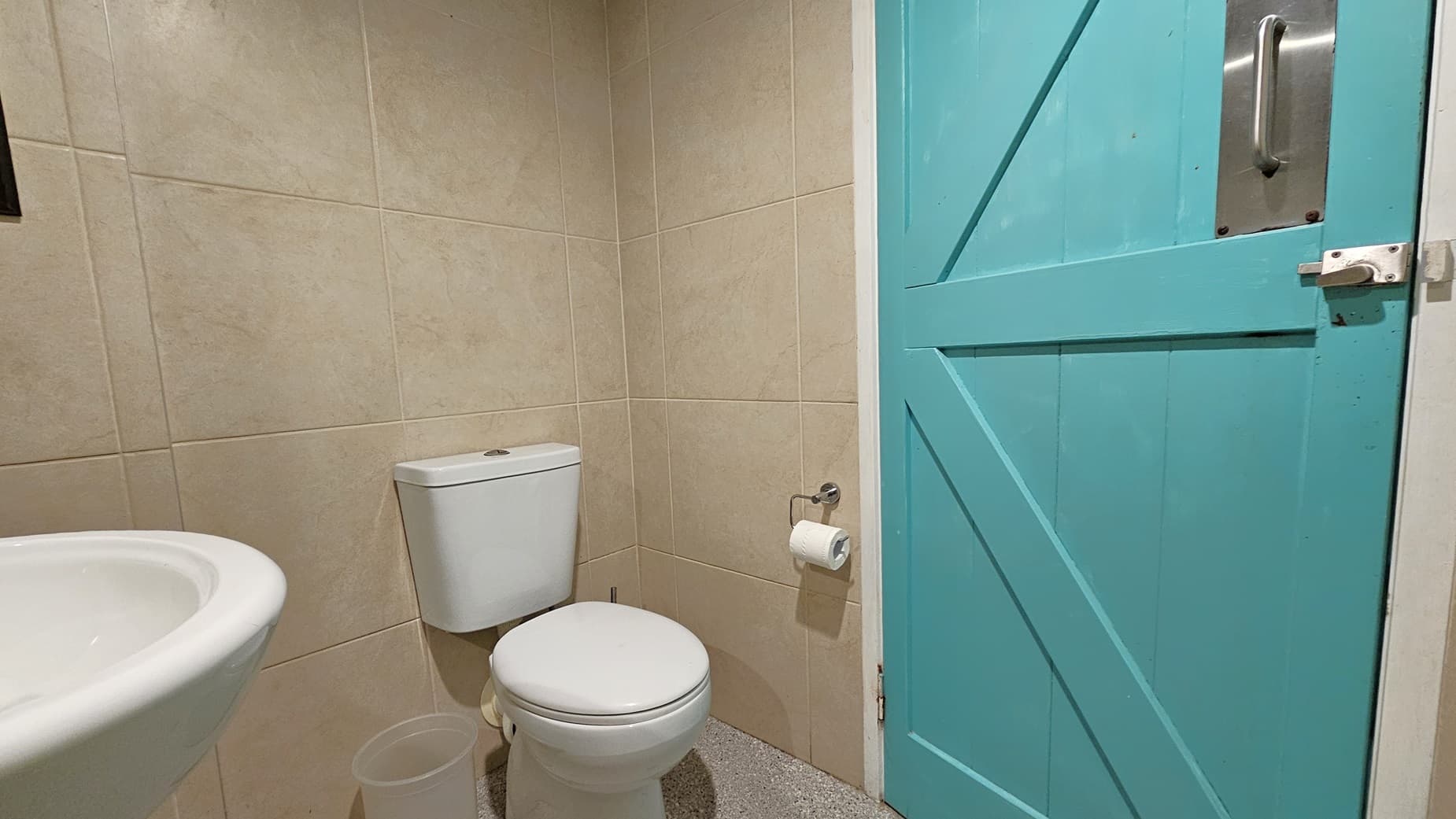 On the left, there is a white sink with a round basin and a beige tiled wall. In the middle, there is a white toilet with a toilet roll holder mounted on the wall beside it. On the right, there is a closed turquoise door with a metal handle and latch. The floor is speckled grey tiles.