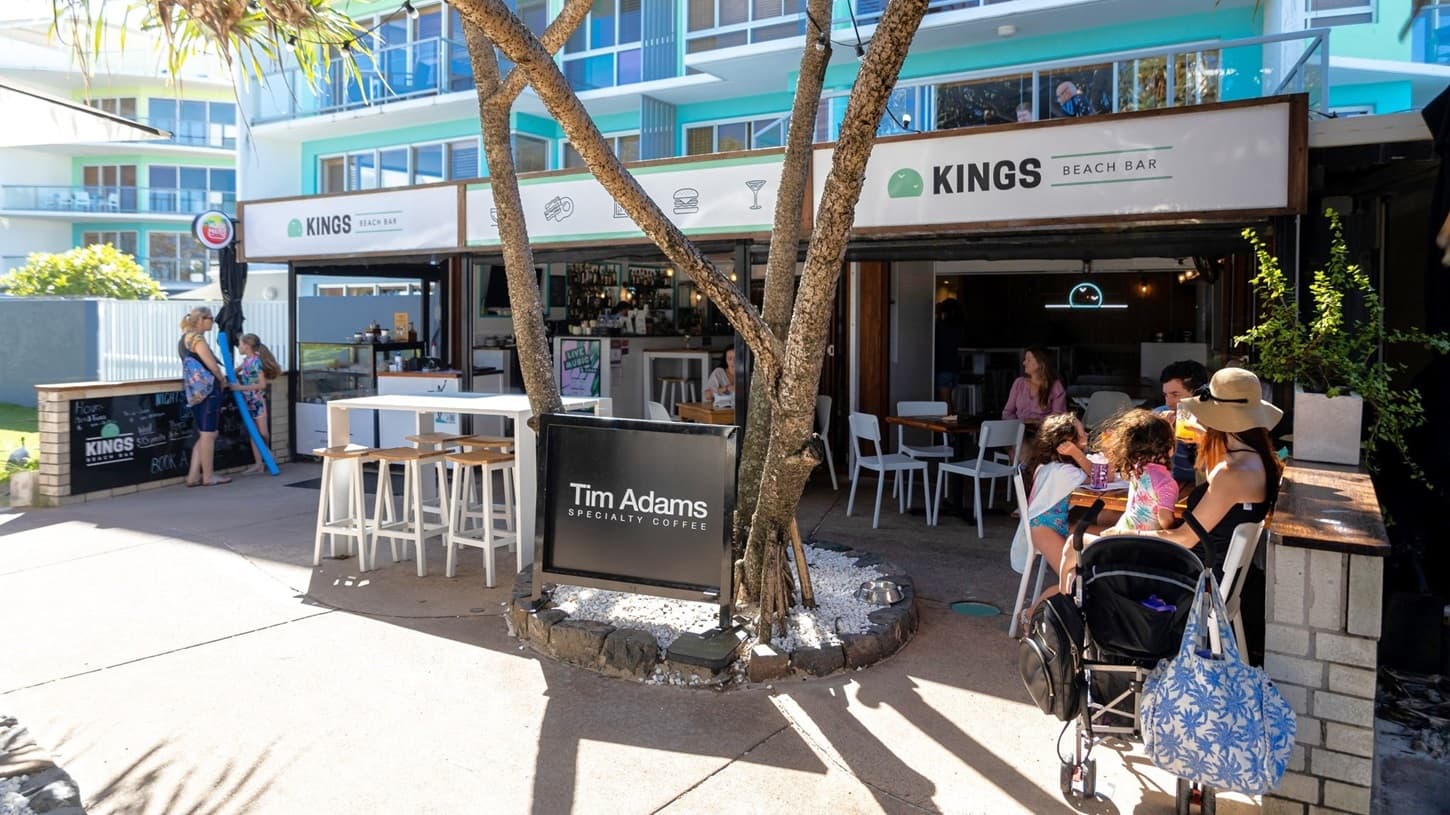 On the left, there is a white fence and a sign for Kings Beach Bar, with people walking past. In the middle, there is an outdoor seating area with white tables and chairs, some occupied by people, and a bar counter with stools. A tall palm tree with a slender trunk  bearing a sign "Tim Adams" stands in the middle, bordered by stones and pebbles. On the right, a family with a pram sits at a table near another smaller tree. The background features a multi-storey building with blue balconies.
