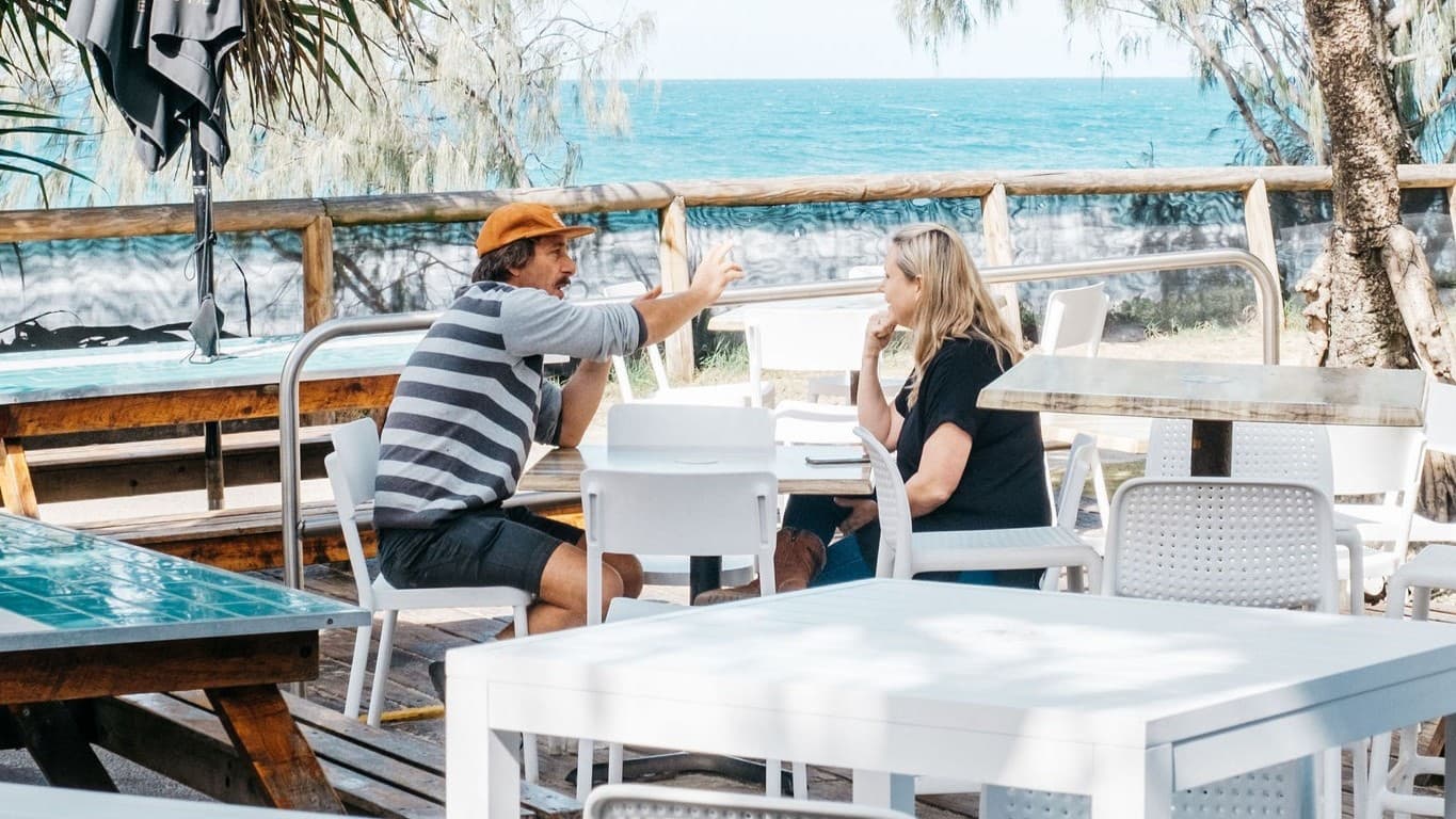There are white tables and chairs with a man in a striped shirt and orange hat sitting and gesturing. In the middle, a woman with blonde hair in a black shirt sits across from him at a white table. On the right, there is a tree with a thick trunk and more white tables and chairs. The background features a wooden fence and the ocean beyond it. The floor is wooden decking, and a folded umbrella shade is positioned on the left.