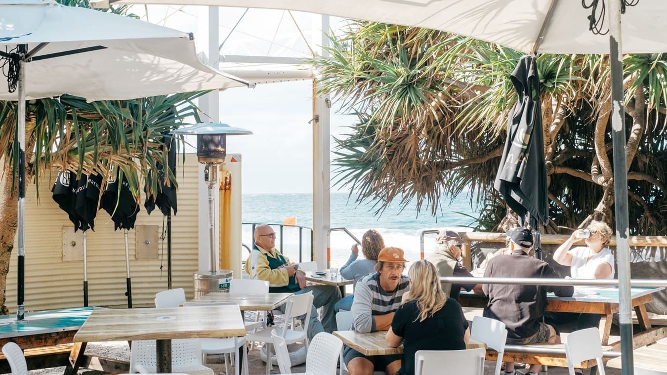 On the left, there are white tables and chairs under a large umbrella, with a man in a yellow jacket sitting at one of the tables. In the middle, a man in an orange hat and a woman with blonde hair sit at a white table, facing each other. On the right, there is a large tree with dense foliage, more tables and chairs, and people sitting and talking. The background features a wooden fence and the ocean beyond it. The floor is wooden decking.