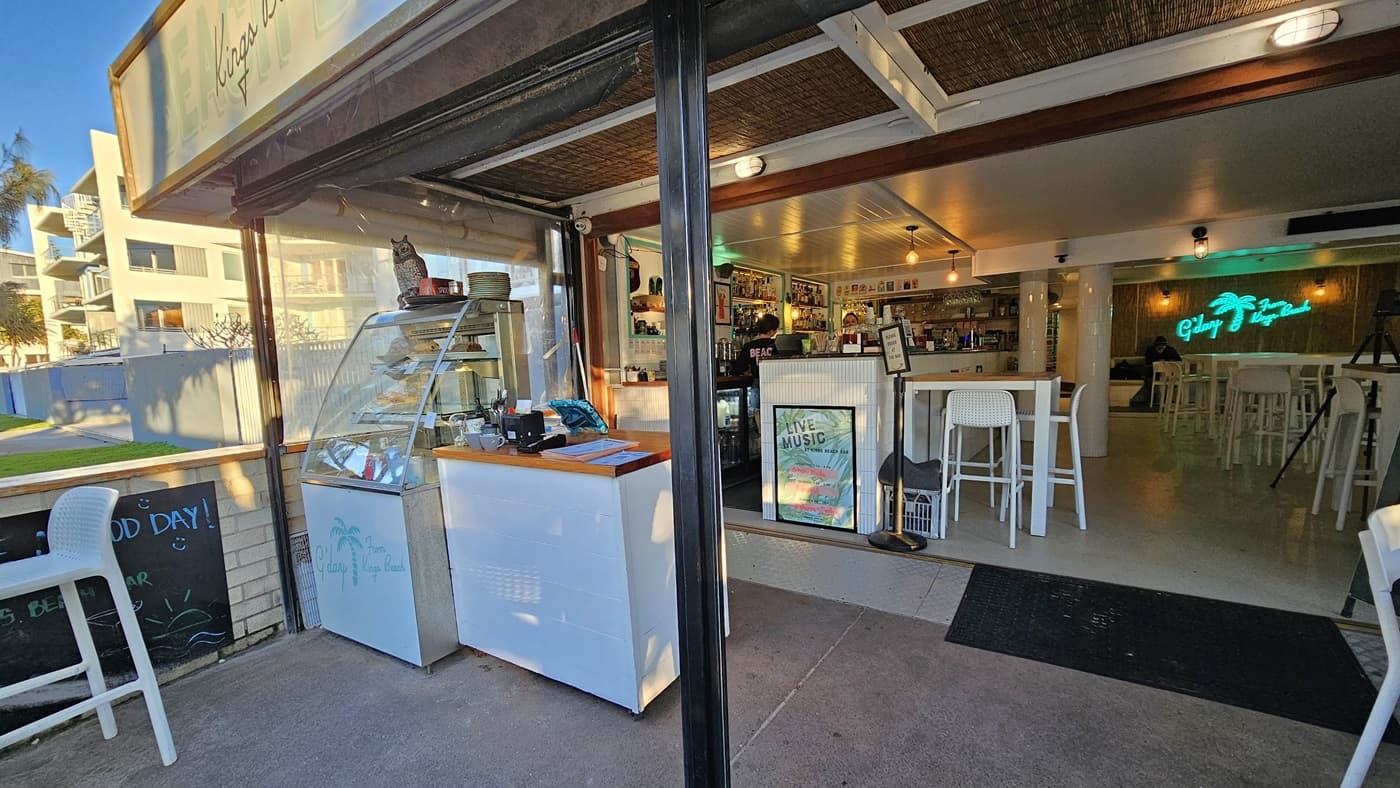 On the left, there is an outdoor area with a white display freezer and a glass display case containing food items. In the middle, there is an open entrance leading into a café with white tables and chairs, a counter with stools, and a neon sign on the back wall. On the right, inside the café, there are more white tables and chairs, and a bar counter with various items. The floor is concrete, and the ceiling has exposed wooden beams and white panels.