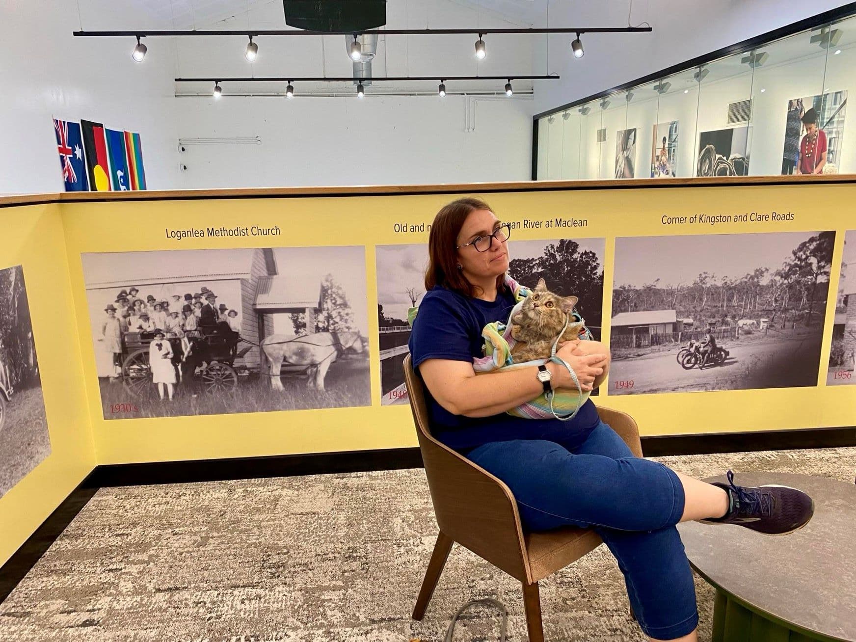 A woman sits in a gallery, holding a small dog before historic photo displays.
