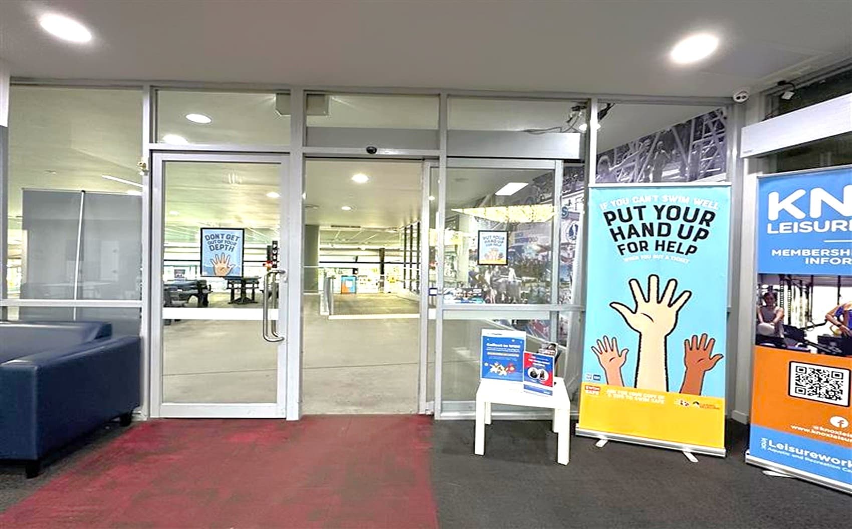 Glass doors in the centre lead to an indoor area with visible gym equipment. A blue and red carpet covers the floor in the foreground. On the right, a large banner with raised hands reads "PUT YOUR HAND UP FOR HELP." Next to it, another banner displays "KN LEISURE" with a QR code. A small white table holds informational leaflets. The ceiling is white with recessed lighting.