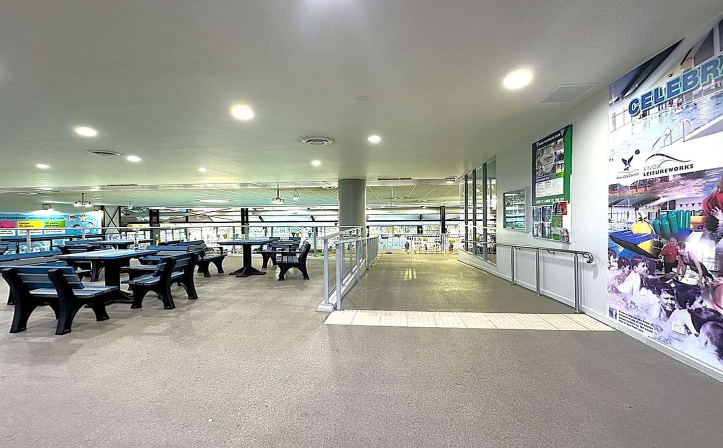A spacious indoor area with a grey carpeted floor and a white ceiling with recessed lighting. On the left, there are several black tables and chairs arranged in rows. The right side features a wall with colourful posters and a railing along a ramp. Large windows in the background provide a view of a swimming pool area. A central column supports the ceiling, and the space is well-lit.