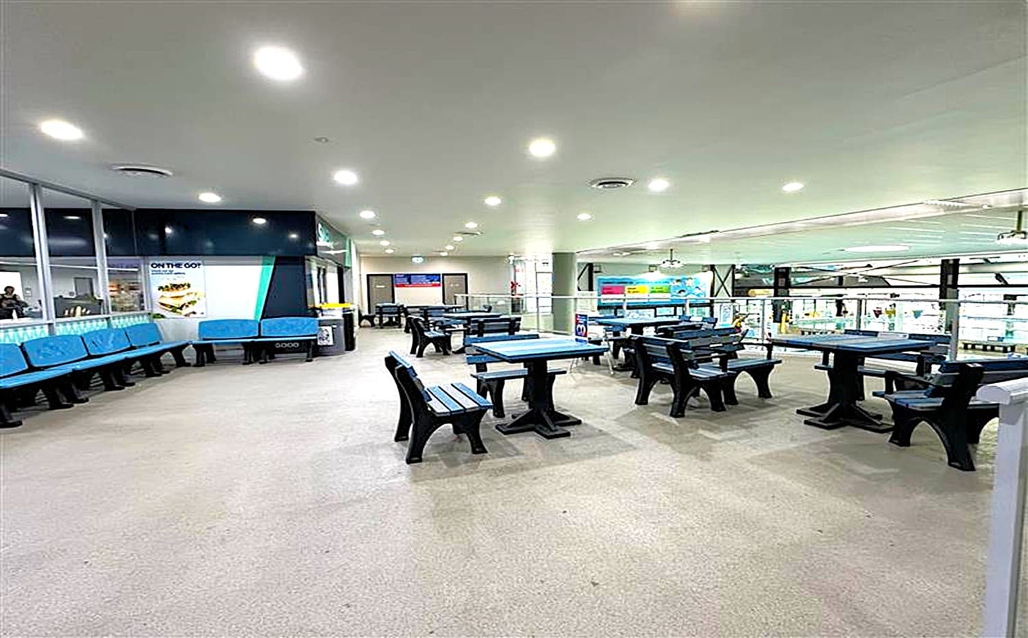 A spacious indoor area with a light-coloured floor and ceiling. On the left, there are blue cushioned benches along a glass wall, with a poster advertising food. In the centre and right, several black and blue picnic-style tables and benches are arranged. The background features a railing overlooking a lower level with bright signage and displays. Ceiling lights are evenly spaced, providing ample illumination.