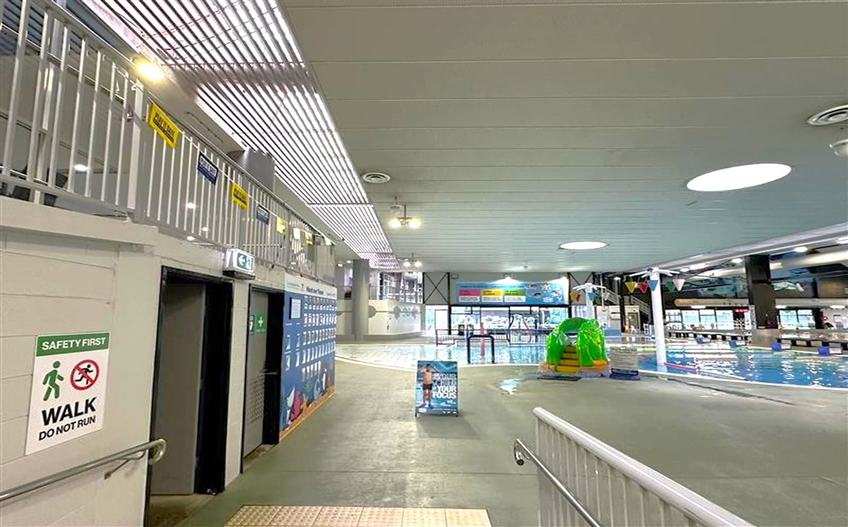Indoor swimming pool area with a grey tiled floor and a white ceiling featuring circular lights. On the left, there are lockers and a sign that reads "SAFETY FIRST WALK DO NOT RUN." A railing runs along the left side. In the centre, a signboard stands on the floor. On the right, a pool with a colourful inflatable structure is visible. The background shows large windows and colourful banners.