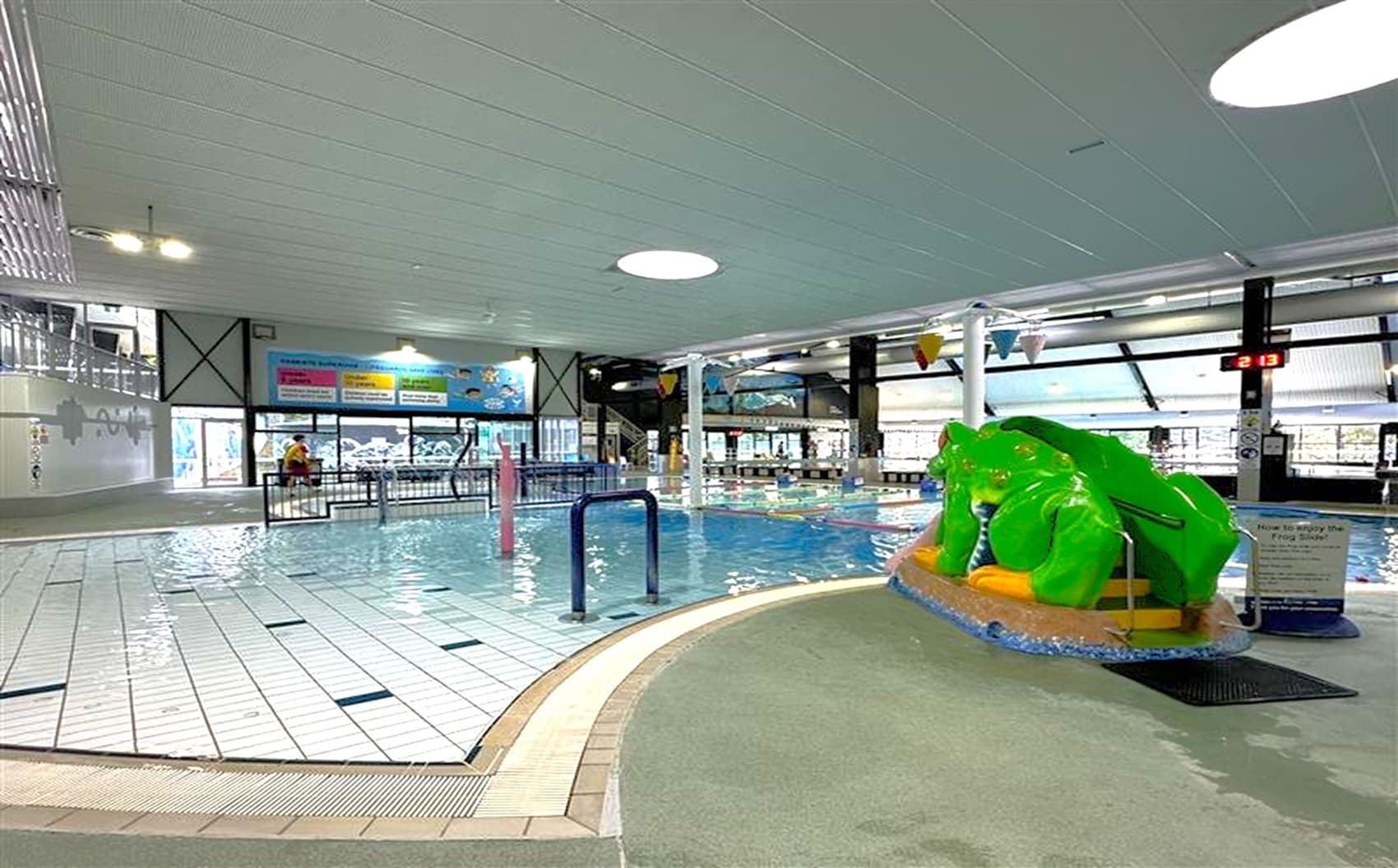 Indoor swimming pool area with a shallow pool on the left, featuring a tiled floor. In the centre, a small slide shaped like a green frog is positioned over the water. The ceiling is white with circular lights. In the background, large windows and colourful banners are visible, along with a digital clock displaying "2:13". A few people are near the entrance, and the area is well-lit with overhead lights.