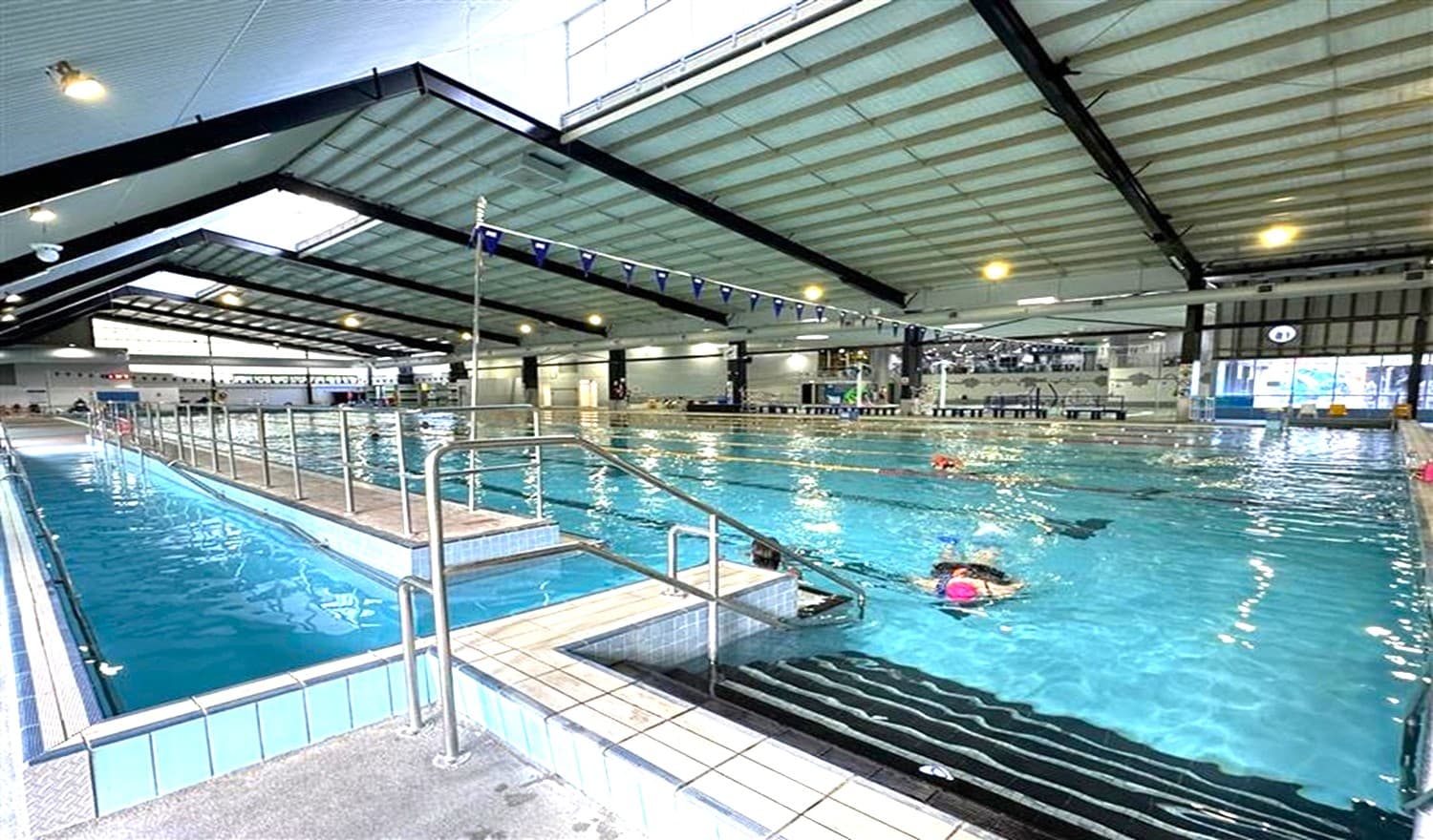 Indoor swimming pool with a high, sloped ceiling featuring exposed beams. On the left, a narrow lane with metal railings and steps leads into the water. The main pool area is on the right, with several swimmers in lanes. The floor is tiled, and the ceiling has large lights. In the background, there are windows and a seating area.