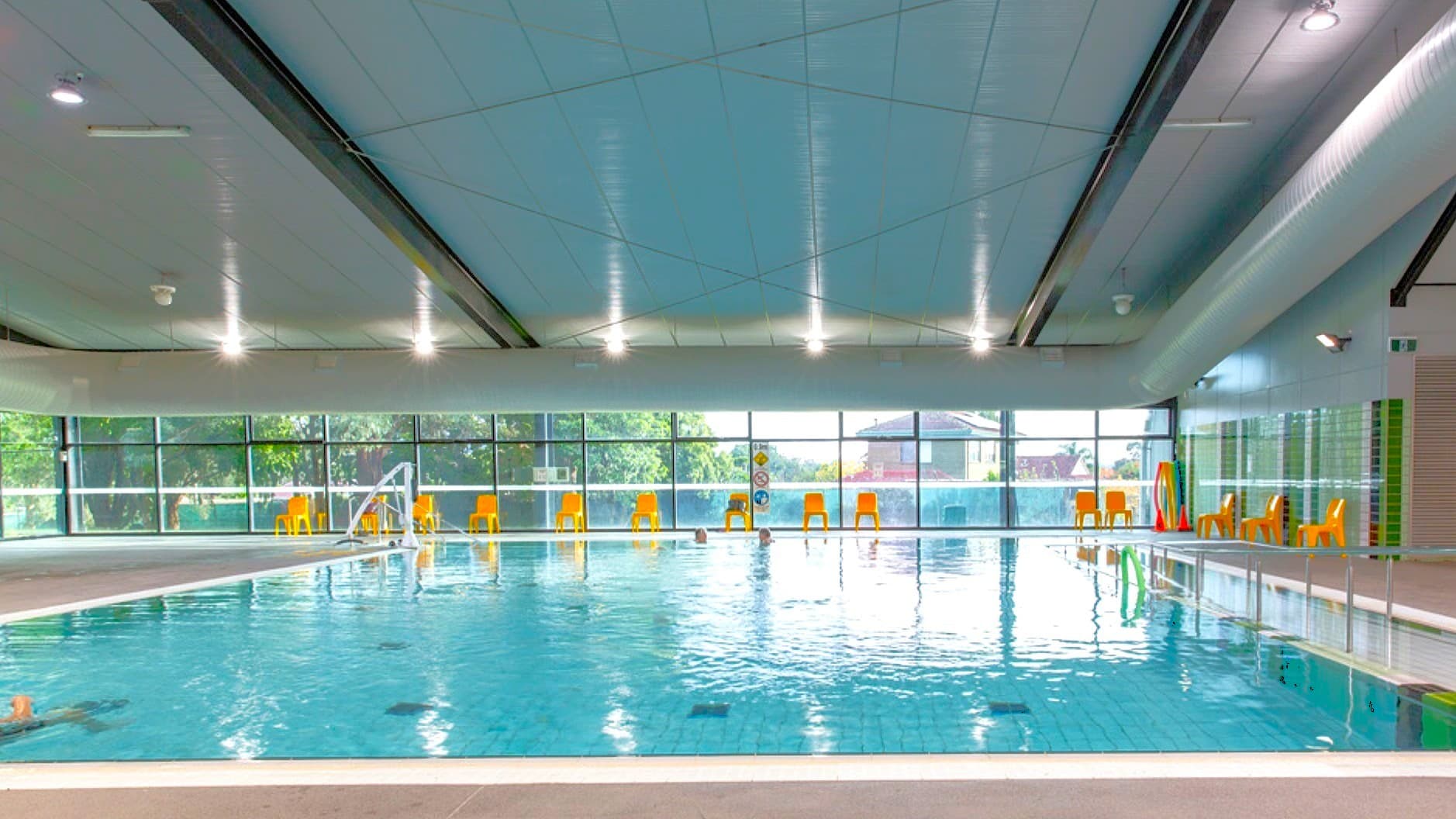 Indoor swimming pool with clear blue water, surrounded by a tiled deck. Several orange chairs are lined up along the glass wall in the background, which offers a view of trees and a building outside. A few people are in the pool. The ceiling is modern with recessed lighting, and the floor is tiled. Bright natural light filters through the large windows.