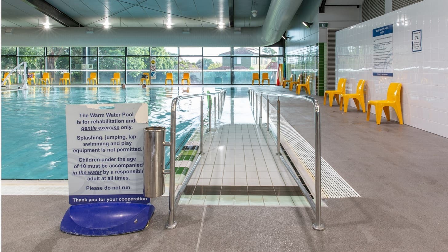 A swimming pool with a ramp leading into the water, bordered by metal railings. A sign at the entrance outlines pool rules, including restrictions on splashing and jumping. On the right, a row of bright yellow chairs lines the tiled wall. Large windows in the background reveal a view of trees and a building outside. The ceiling is equipped with lighting, and the floor is tiled in a light colour.