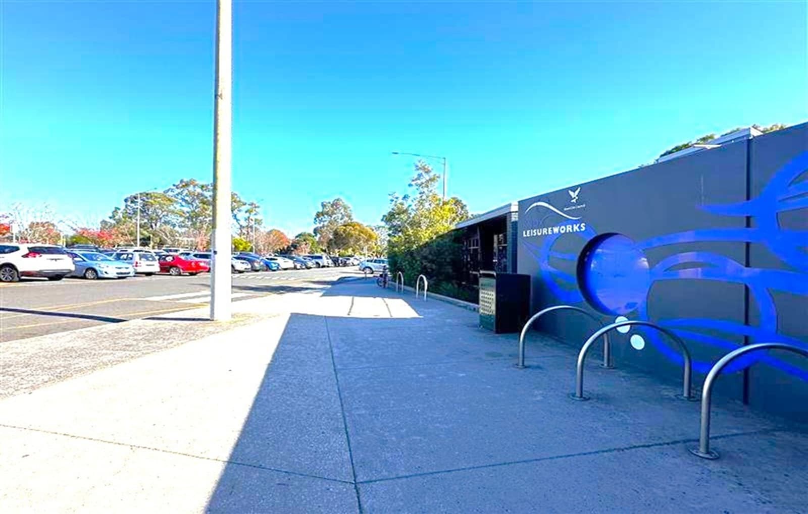 A paved walkway runs alongside a construction site on the right, with a dark blue hoarding displaying the words "Future Works." Metal bike racks are positioned in front of the hoarding. On the left, a car park is visible with several parked vehicles, including white and red cars. A tall streetlight stands near the walkway. In the background, there are trees and a clear blue sky. The ground is a mix of concrete and asphalt.