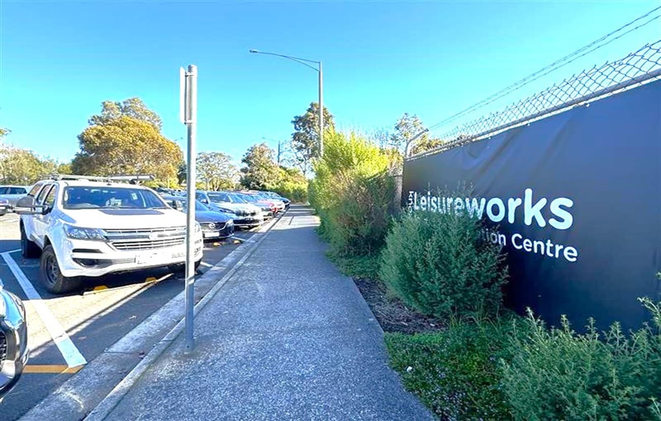 A paved footpath runs through the centre, bordered by a row of parked cars on the left, including a white SUV. On the right, a tall fence with barbed wire tops a sign that reads "Leisureworks Recreation Centre." Bushes and small trees line the fence. In the background, larger trees are visible under a clear blue sky. A metal pole stands near the path.