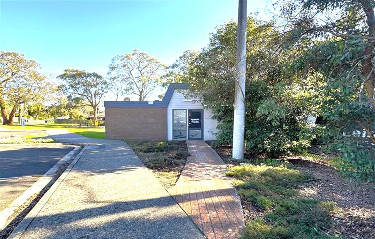 A small, single-storey brick building is situated in the centre, with a glass door and a sign above it. A narrow brick path leads to the entrance, flanked by low shrubs. On the right, a tall tree and a utility pole are visible. The left side features a paved road and more trees in the background. The area is well-lit, suggesting daytime.