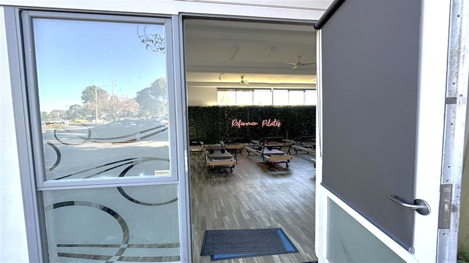A partially open door reveals an interior space with wooden flooring. Inside, several Pilates reformer machines are arranged in rows. A sign on the back wall reads "Reformer Pilates" in illuminated letters. The room has large windows allowing natural light to enter. The door on the left has a glass panel with decorative circular patterns, and outside, trees and a road are visible. The ceiling inside is plain with light fixtures.