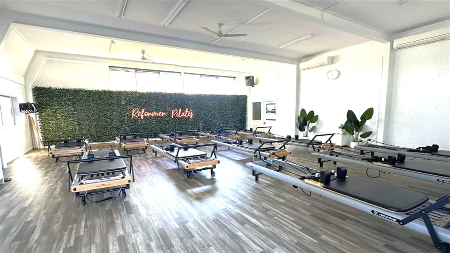 A Pilates studio with several reformer machines arranged in rows on a wooden floor. The back wall features a green plant wall with a neon sign reading "Reformer Pilates". Large windows above the plant wall allow natural light to enter. On the right, there are potted plants and a wall clock. The ceiling is white with a fan in the centre.