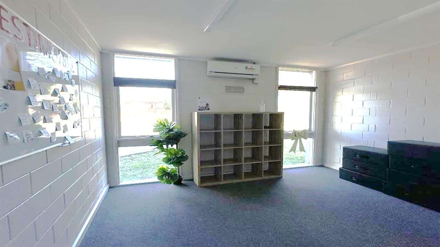 A room with two tall windows on the back wall, allowing natural light to enter. In the centre, there is a tall shelving unit. To the left, a wall displays a board with various papers or notes attached. A potted plant is positioned near the left window. The floor is carpeted, and the ceiling is plain with visible light fixtures. An air conditioning unit is mounted above the shelving unit.