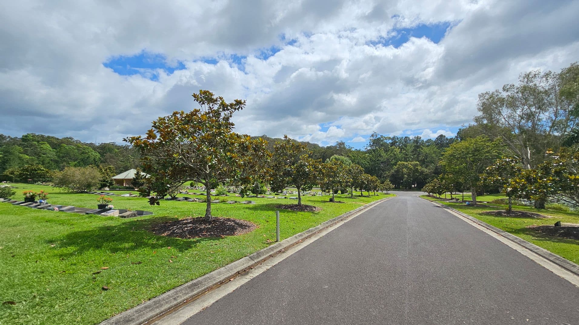 A paved road runs through the centre, flanked by neatly trimmed grass and rows of small trees. On the left, there are gravestones and flower arrangements scattered across the lawn. A small building with a sloped roof is visible in the background. On the right, more trees line the road, with dense forest in the distance. The scene is calm and orderly, with a mix of open space and greenery.