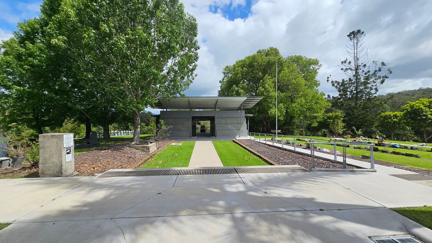 A pathway leads to a modern, open-sided structure with a flat roof, surrounded by lush green trees. On the left, a concrete post with signage stands near a tree-lined area with gravestones. On the right, a manicured lawn with a row of low plants and a metal railing. The background features more trees and a slightly overcast sky. The foreground shows a concrete pavement with shadows from the trees.