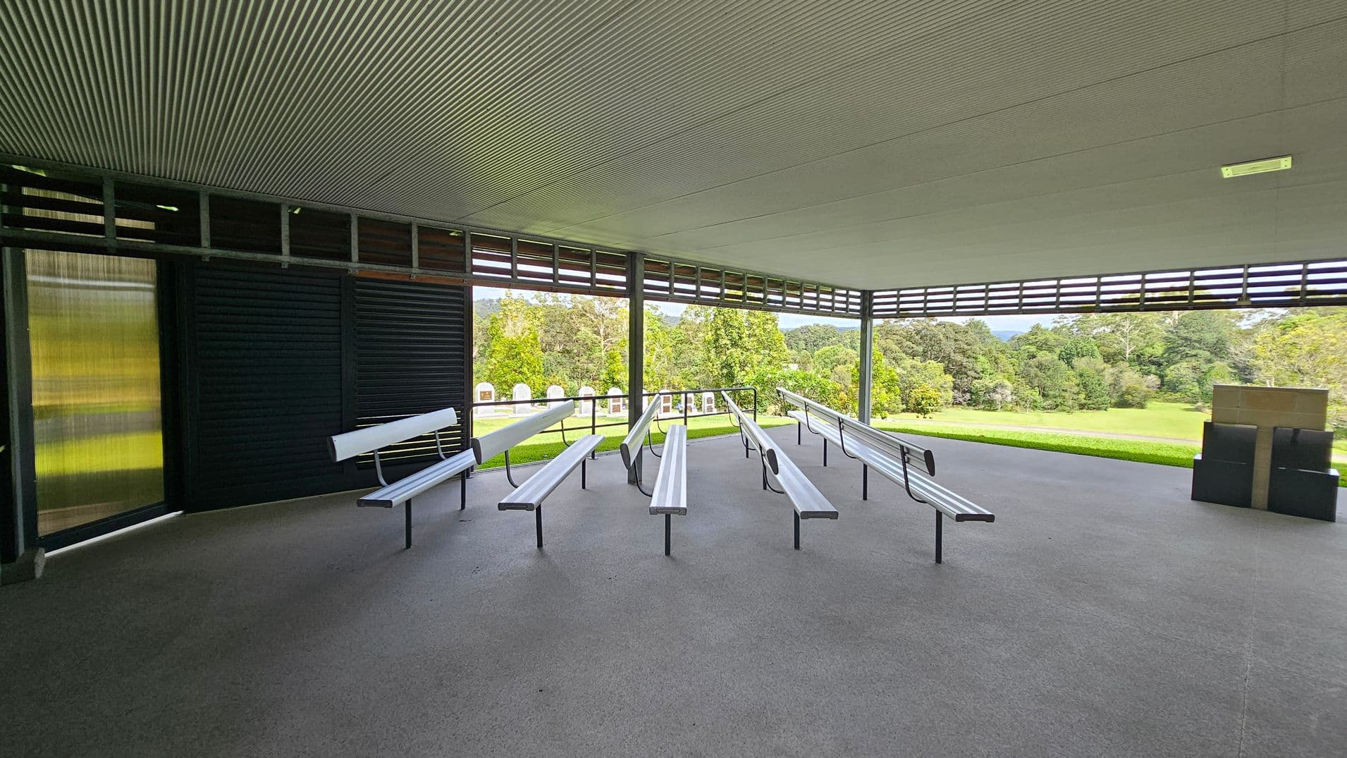 An open-air pavilion with a corrugated ceiling and grey floor. In the centre, there are several rows of metal benches facing forward. The left side has a dark louvred wall, while the right side opens to a grassy area with trees in the background. A stack of black boxes is on the right. The pavilion is surrounded by a lush, green landscape.