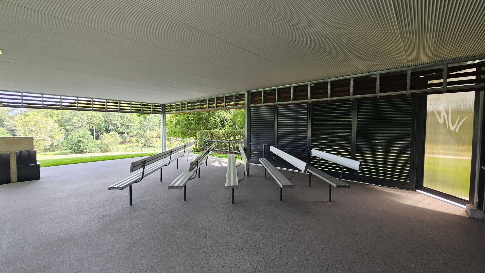 An open-air pavilion with a grey, textured floor and a ribbed ceiling. In the centre, there are several metal benches with backrests arranged in rows. The right side features dark louvred panels, while the left side is open, offering a view of lush green trees and grass. A small structure with a flat top is visible on the far left. Natural light filters through the space, creating a serene atmosphere.