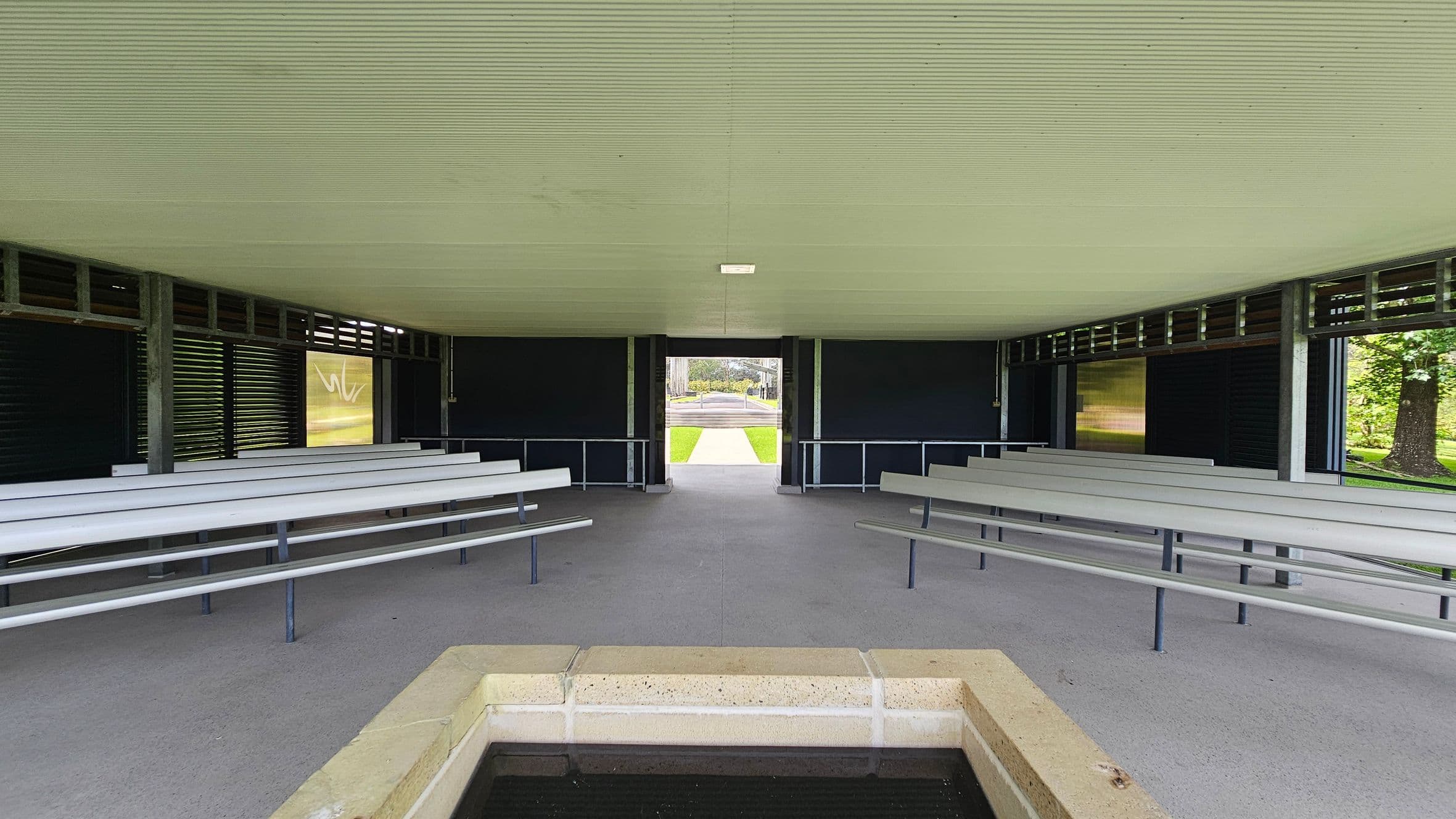 A covered outdoor pavilion with a corrugated ceiling and concrete floor. In the foreground, there is a rectangular stone structure, possibly a water feature. On both sides, there are rows of grey benches facing inward. The background shows an open exit leading to a grassy area and a path. The walls have dark panels with horizontal slats, allowing some light to filter through. Trees are visible outside on the right.