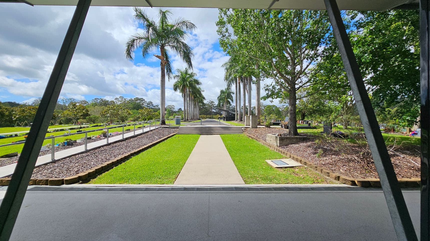 A pathway leads through a garden with neatly trimmed grass, flanked by tall palm trees on both sides. On the left, a railing runs alongside a gravel path, with more greenery and distant trees. On the right, a large tree stands in a raised bed with mulch. In the background, a structure is partially visible, surrounded by more trees. The foreground shows a covered area with a grey floor and metal beams.