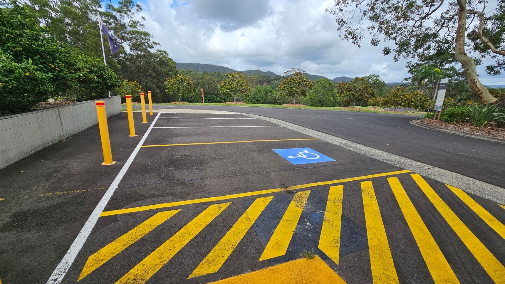 A parking area with a designated disabled parking space marked by a blue symbol on the ground. Yellow diagonal lines are painted on the asphalt in the foreground. On the left, there are several yellow bollards and a low concrete wall with greenery behind it. The background features lush trees and hills under a cloudy sky. On the right, a sign is visible near more vegetation. The area is surrounded by natural scenery.