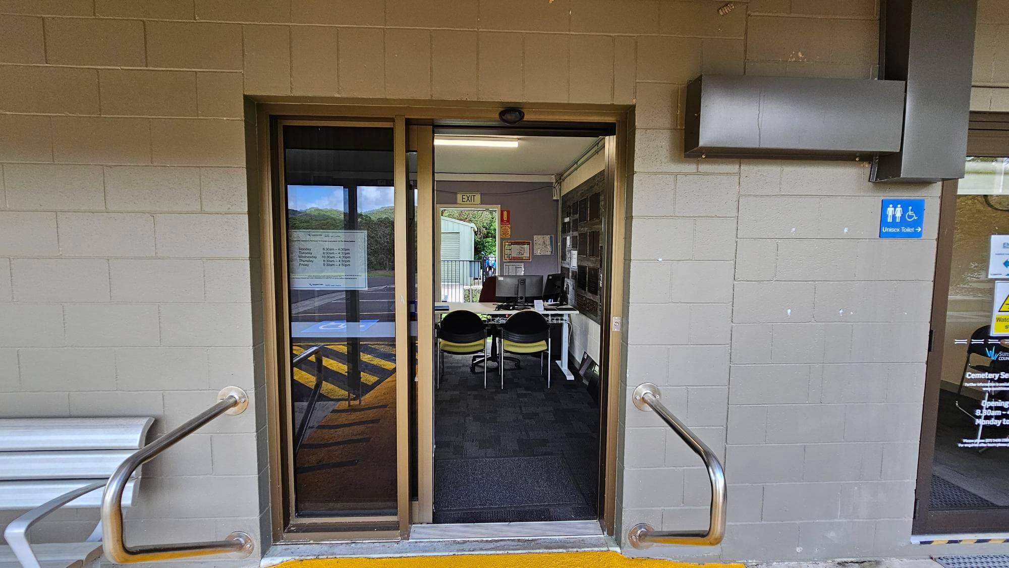 A doorway with a sliding glass door leads into an office space. Inside, there are two chairs facing a desk with a computer. The walls are made of light-coloured bricks. On the right, a sign indicates the direction to a unisex toilet. Metal handrails are on either side of the entrance. The floor outside is marked with yellow paint, and the interior has dark carpeting. An "EXIT" sign is visible inside above the door.
