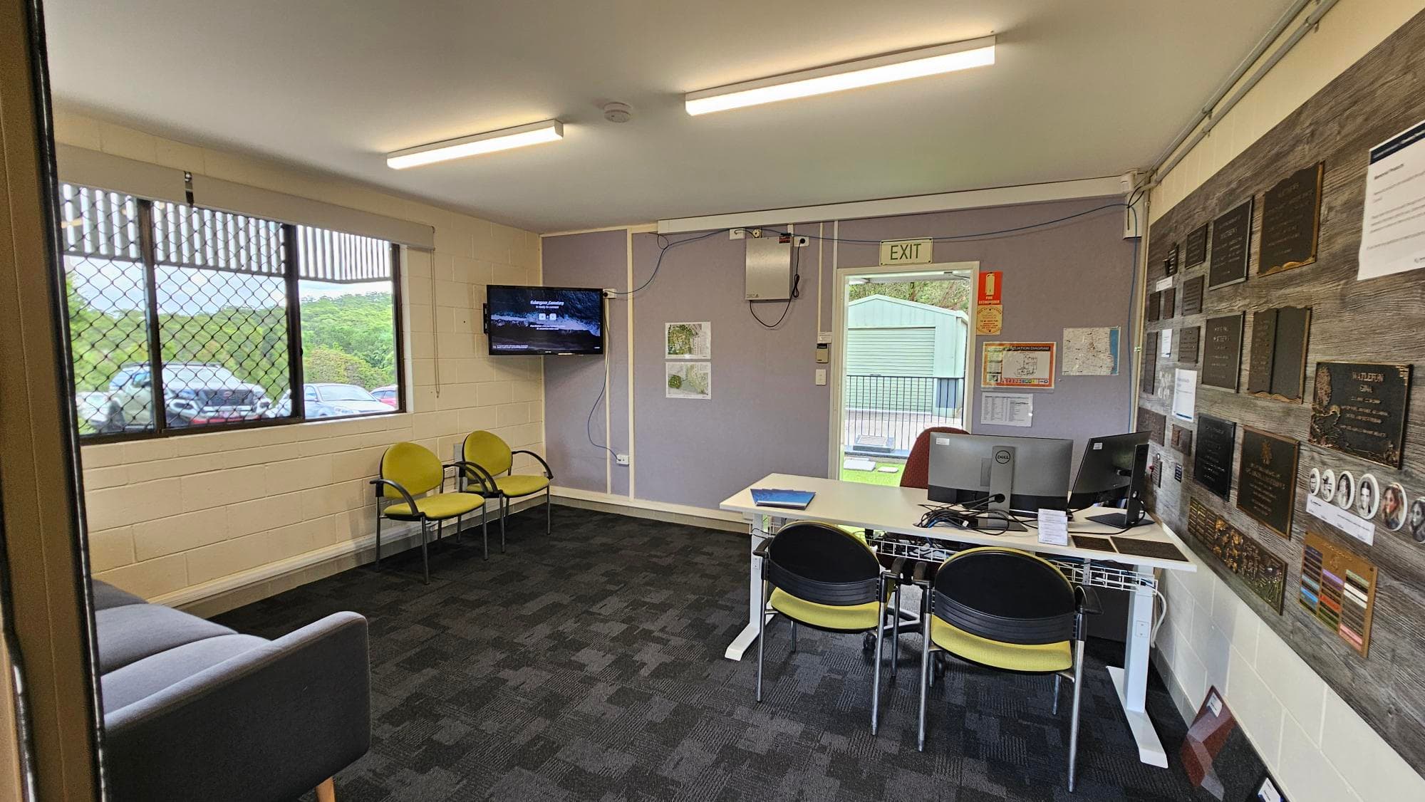 A room with a dark carpeted floor and fluorescent lights on the ceiling. On the left, there are windows with metal grilles and a view of parked cars outside. Below the windows, a grey sofa and two yellow chairs are placed. In the middle, a wall-mounted TV is visible. On the right, a desk with two computer monitors and chairs is positioned. The wall behind the desk is covered with plaques and notices. An open door leads outside, showing a green building in the background.
