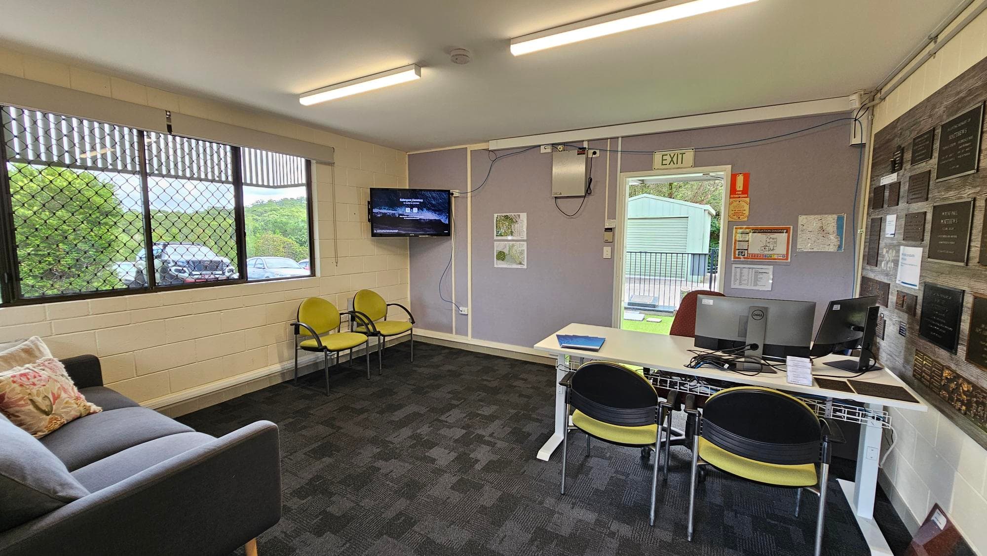 A small office space with a grey sofa and colourful cushions on the left, next to a large window with a metal grid. Two yellow chairs are positioned near the window. In the middle, a wall-mounted TV is visible. On the right, a desk with two computer monitors and chairs faces a wall adorned with plaques. An exit door is nearby, leading outside. The floor is carpeted in a dark pattern, and the ceiling has fluorescent lights.