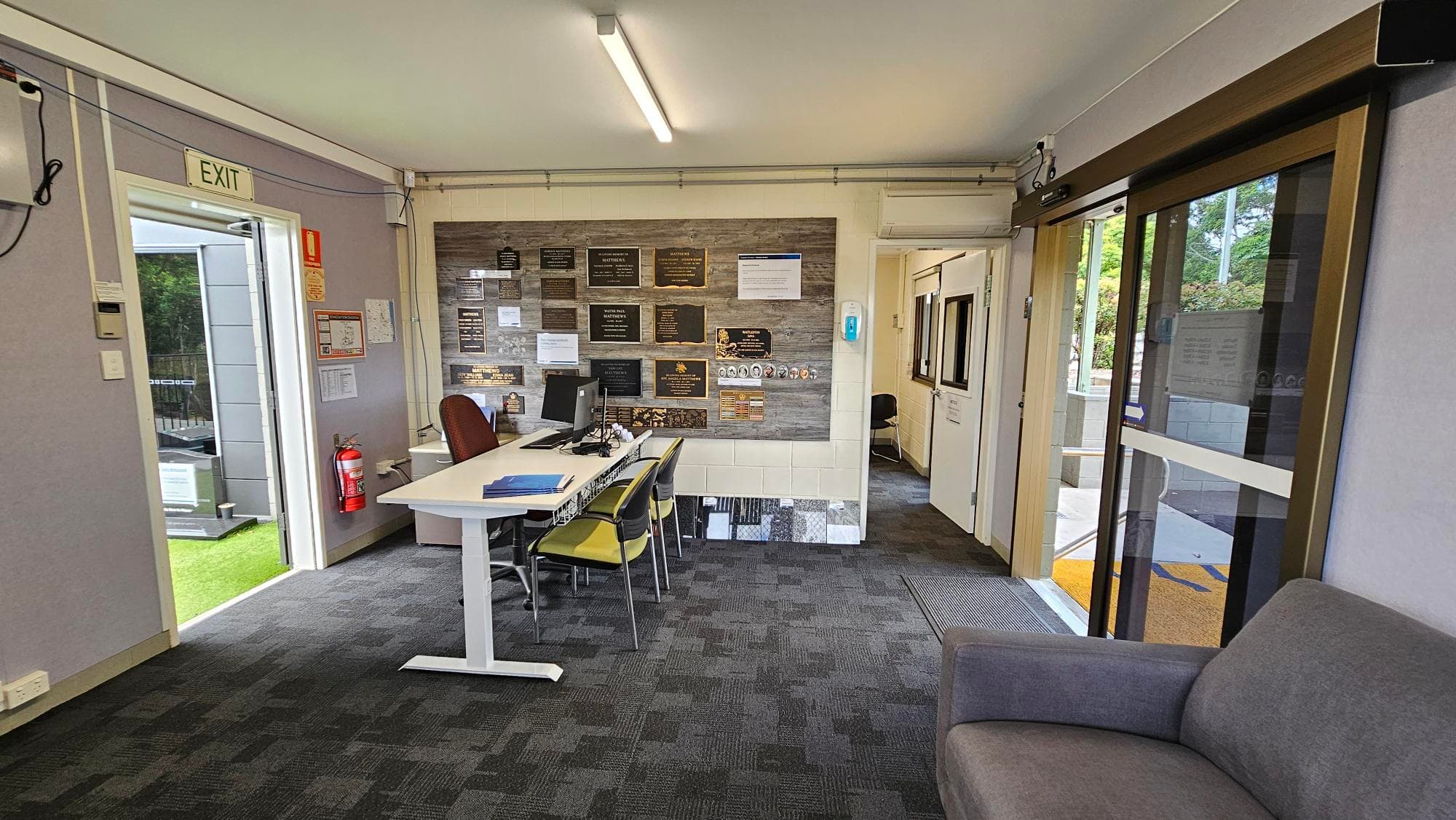 A small office space with a grey carpeted floor. On the left, there's an open door with an "EXIT" sign above it and a fire extinguisher nearby. In the centre, a white desk with a computer and chairs faces a wall adorned with various plaques and notices. On the right, a glass sliding door leads outside, and a grey sofa is partially visible. The ceiling has a fluorescent light fixture.