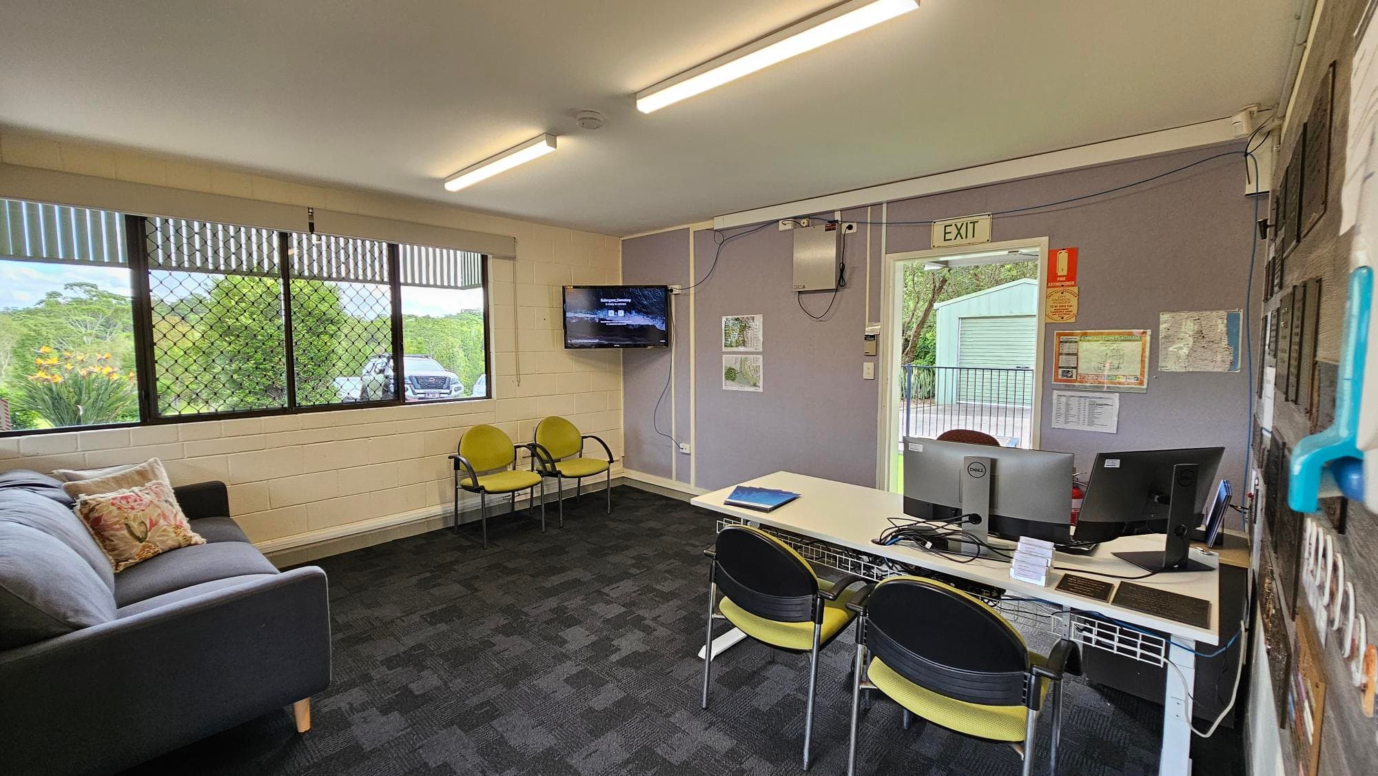 A room with a grey sofa and decorative cushions on the left, next to a window with a view of greenery and a parked car outside. In the middle, there are two yellow chairs and a wall-mounted TV. On the right, a desk with two computer monitors and chairs. The wall has maps and an exit sign above a door leading outside. The floor is carpeted in dark grey, and the ceiling has fluorescent lights.