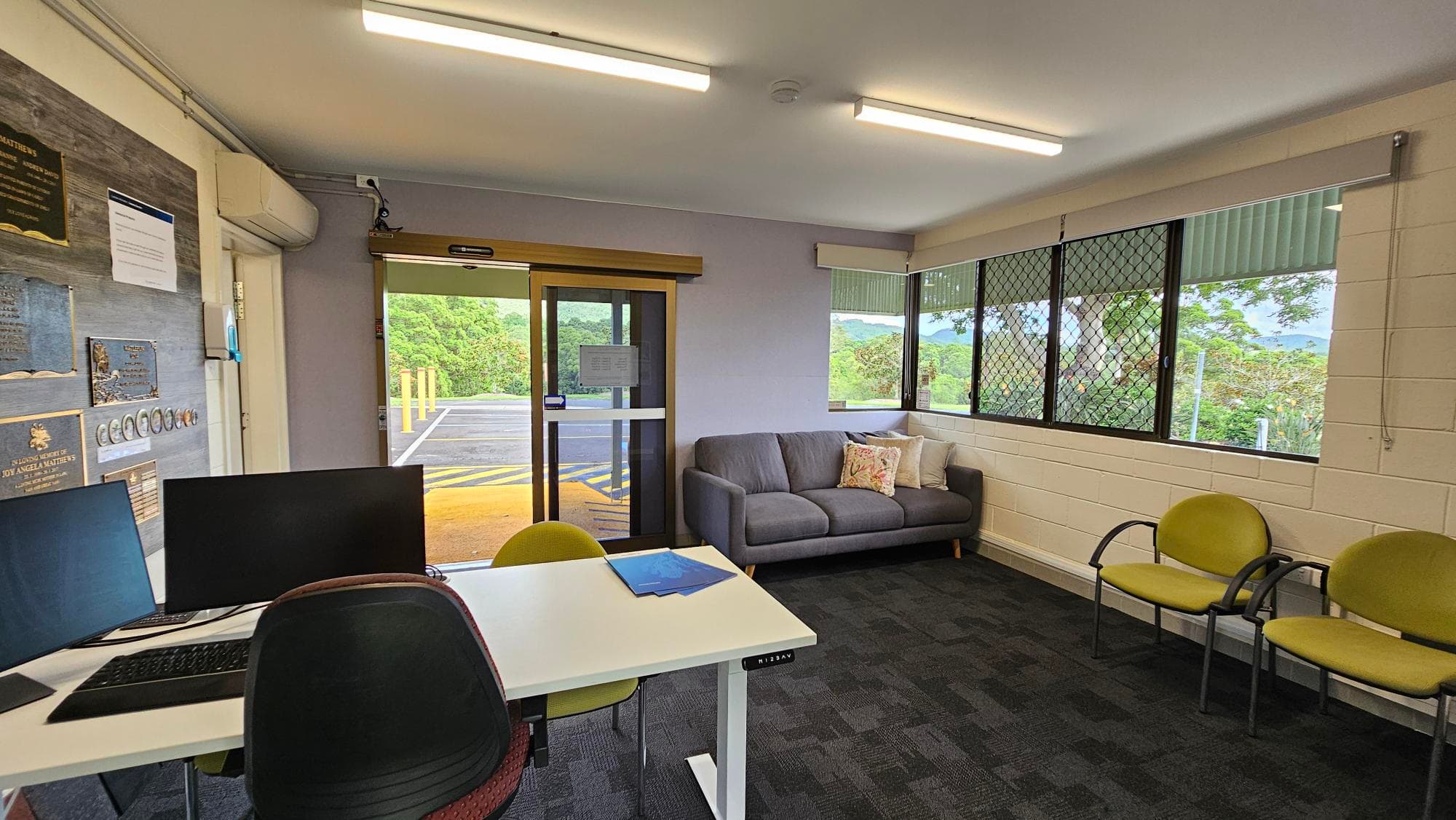A small office space with a desk on the left featuring two computer monitors and a chair. The wall behind the desk has several plaques and notices. In the middle, a glass door leads outside, allowing natural light in. On the right, a grey sofa with decorative cushions is placed against a wall with large windows, offering a view of greenery outside. Two green chairs are positioned near the sofa. The floor is carpeted, and the ceiling has fluorescent lights.