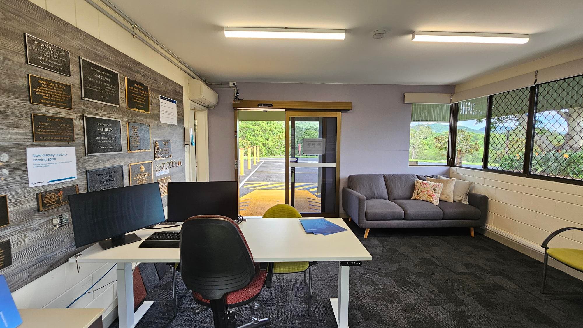 Office interior with a desk and two chairs in the foreground, featuring two computer monitors. The left wall displays numerous plaques. In the background, a glass door leads outside, and a grey sofa with cushions is positioned against the right wall. Large windows with mesh screens provide a view of greenery outside. The floor is carpeted in a dark pattern, and the ceiling has fluorescent lighting.