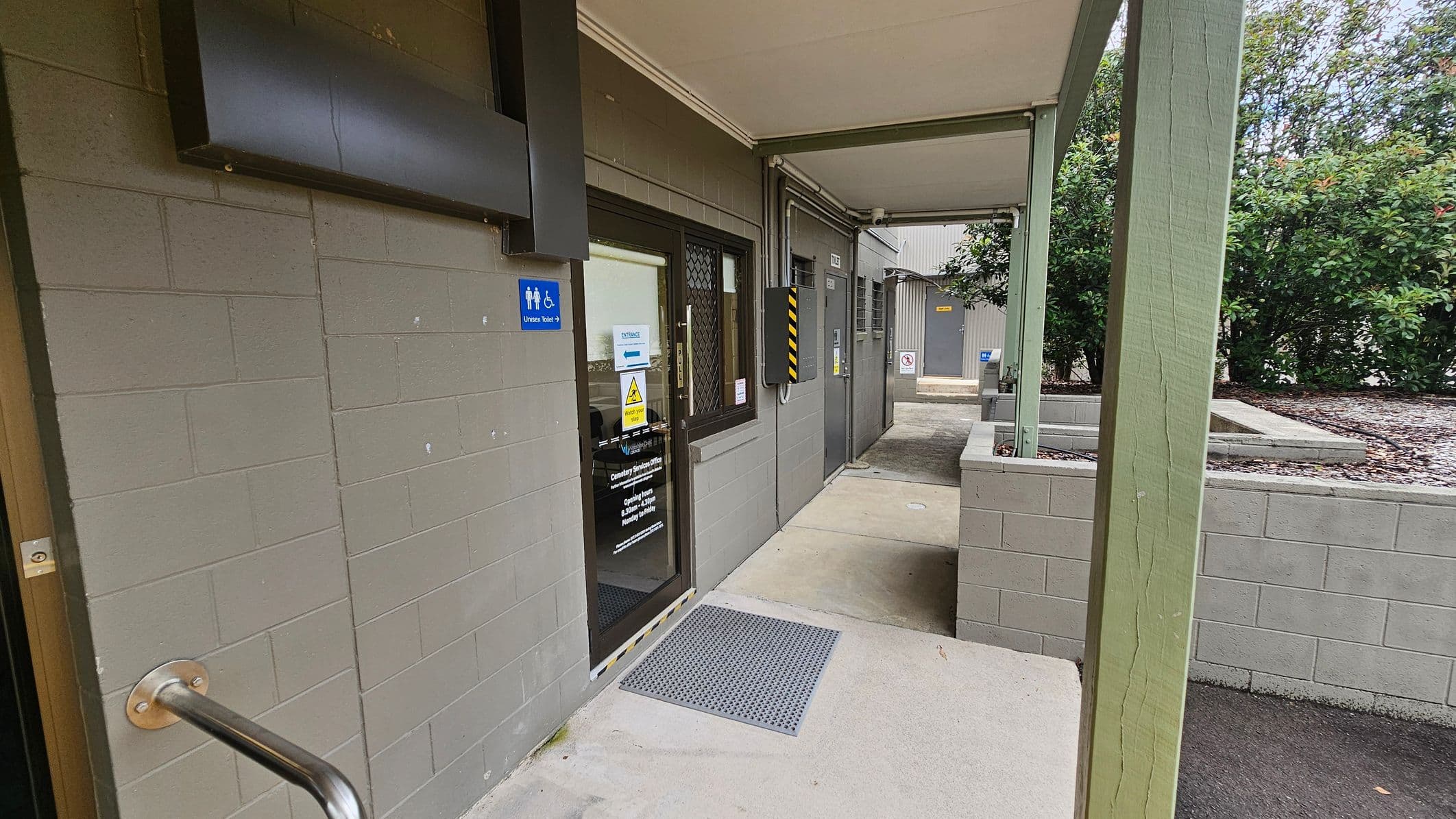 A covered walkway with a green-painted brick wall on the left, featuring a door with a glass panel and a sign indicating a universal toilet. A metal handrail is attached to the wall. The floor is concrete with a textured mat near the door. On the right, a green post supports the roof, and there are bushes in the background. Further along the walkway, there are additional doors and a small courtyard area.