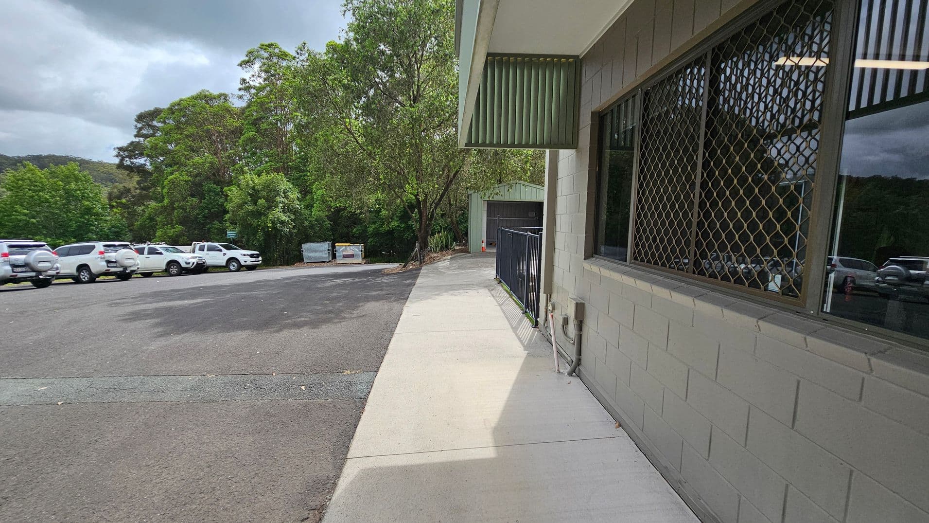 On the right, there's a building with a grey brick wall and a window covered by a metal grid. A narrow concrete path runs alongside the building. On the left, several white vehicles are parked on an asphalt surface. In the background, there are dense green trees and a couple of large bins. The sky is overcast, casting a muted light over the scene.