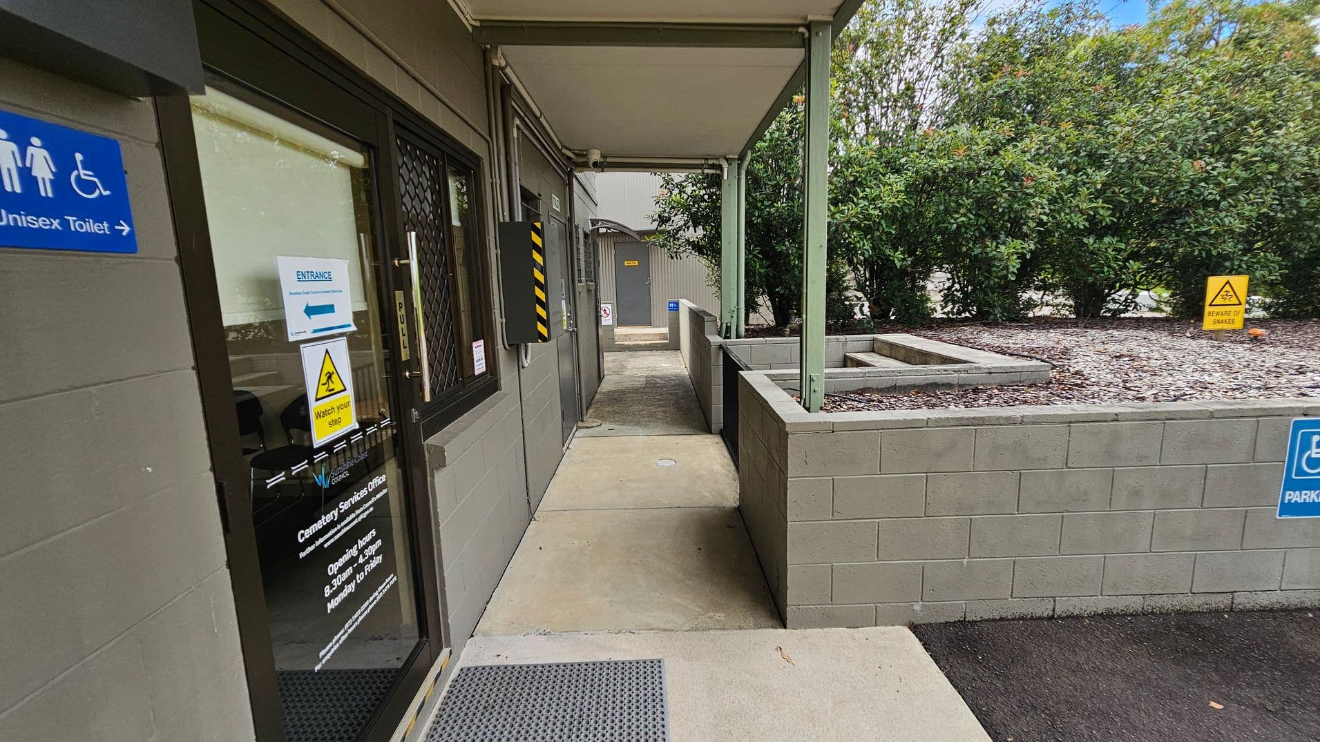 On the left, there is a glass door with signage indicating a unisex toilet and entrance information. The door has a "Watch your step" warning. In the middle, a covered walkway leads to another door in the distance. On the right, a low wall with a garden bed filled with gravel and a "Beware of snakes" sign. Bushes are visible in the background. The floor is concrete, and the ceiling of the walkway is flat and supported by green metal beams.