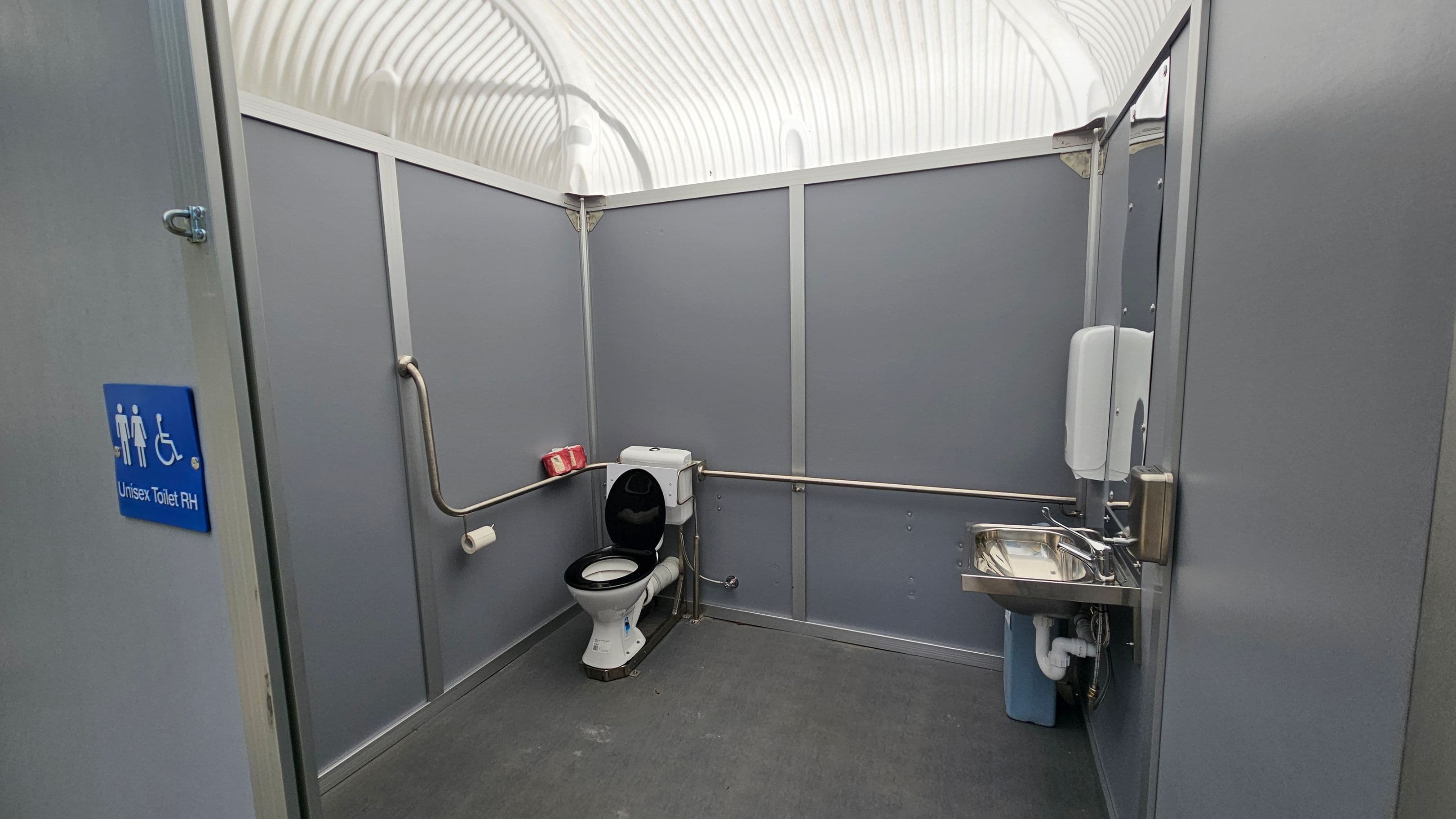 A grey-walled unisex toilet with accessibility features. On the left, a blue sign indicates "Unisex Toilet RH". In the middle, a toilet with a black seat is positioned against the wall, with a grab rail beside it. A roll of toilet paper is mounted on the wall. On the right, a stainless steel sink with a soap dispenser above it. The ceiling is arched and translucent, allowing light to filter through. The floor is grey and appears to be made of a non-slip material.