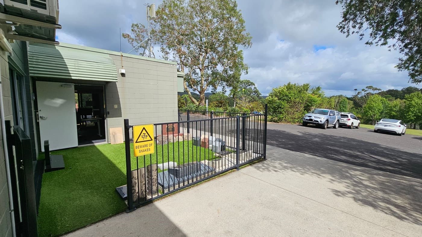 On the left, there's a building with a light green exterior and an open white door. A small area of artificial grass is enclosed by a black metal fence with a "Beware of Snakes" sign. In the middle, a tree is visible behind the building. On the right, a paved area with three parked cars is surrounded by greenery. The ground is a mix of concrete and asphalt.