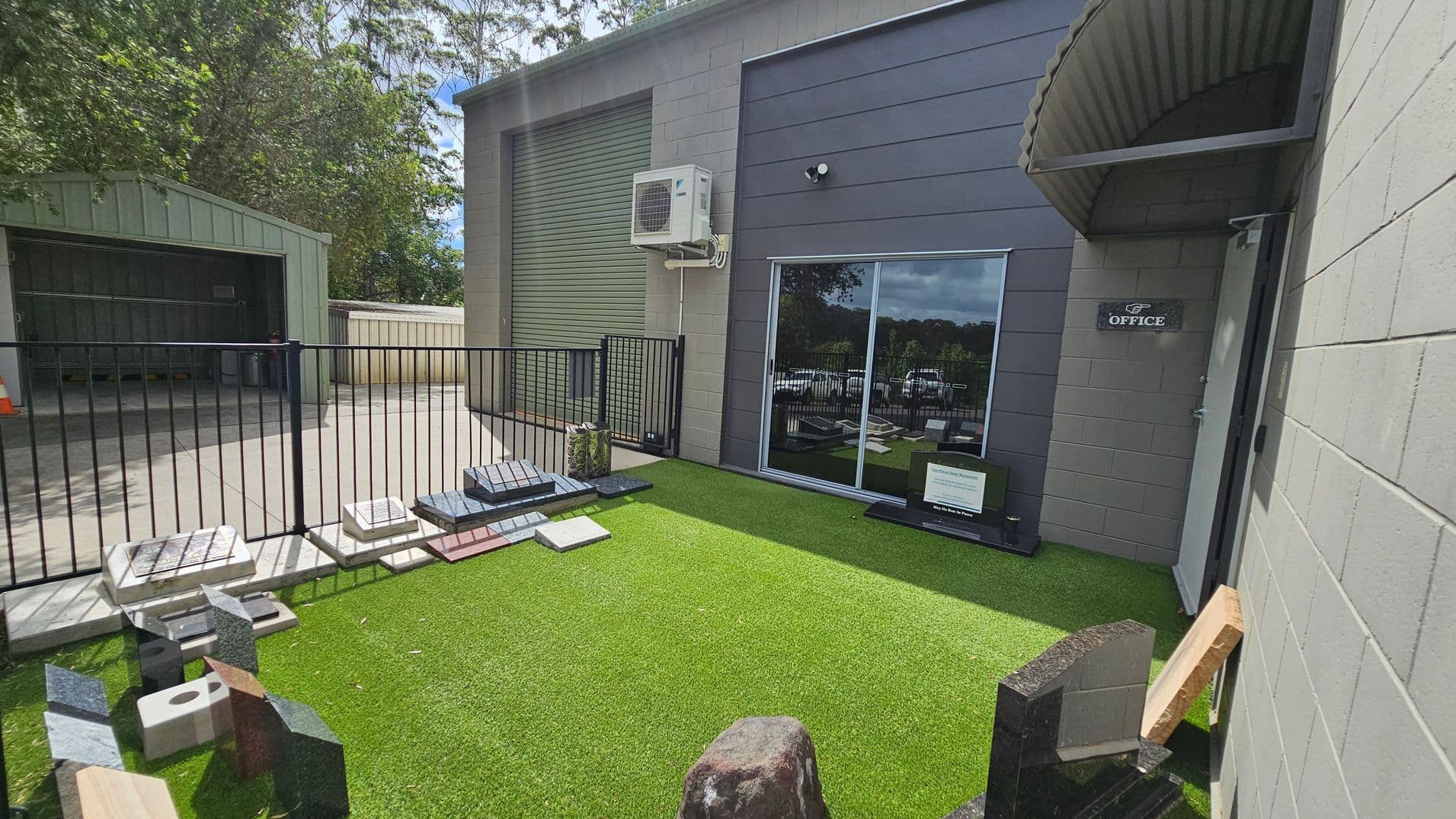 A small outdoor area with artificial grass on the ground. On the left, there is a metal fence with various stone samples displayed along its base. Behind the fence, a shed with an open door is visible. On the right, a building with a grey exterior features a sliding glass door and a sign marked "Office" next to it. An air conditioning unit is mounted above the door. Trees are visible in the background.
