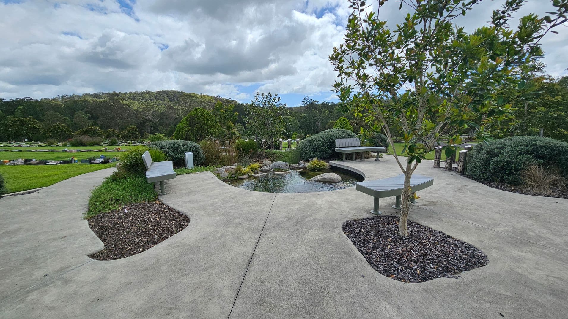 A landscaped garden with a concrete path winding through the centre. In the middle, there's a small tree surrounded by a circular patch of mulch. Benches are placed along the path, with shrubs and bushes neatly arranged around them. The background features a dense line of trees, and the grass is well-maintained. The sky is overcast with clouds.