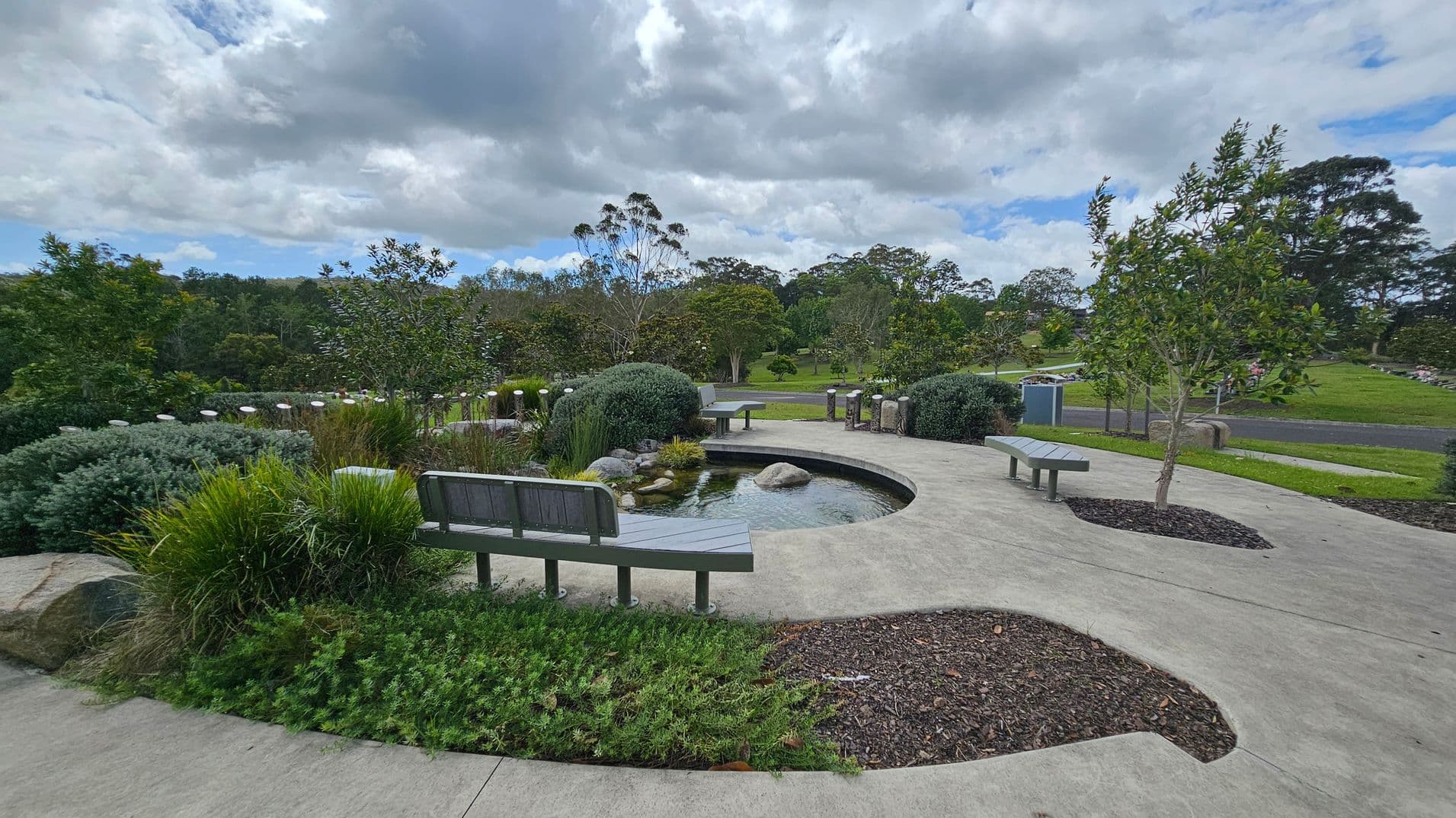 A landscaped garden with a curved concrete path and a small pond in the centre, surrounded by rocks and lush greenery. Wooden benches are placed along the path, offering seating. Trees and shrubs are scattered throughout the area, with a grassy field and more trees in the background. The sky is overcast with thick clouds.