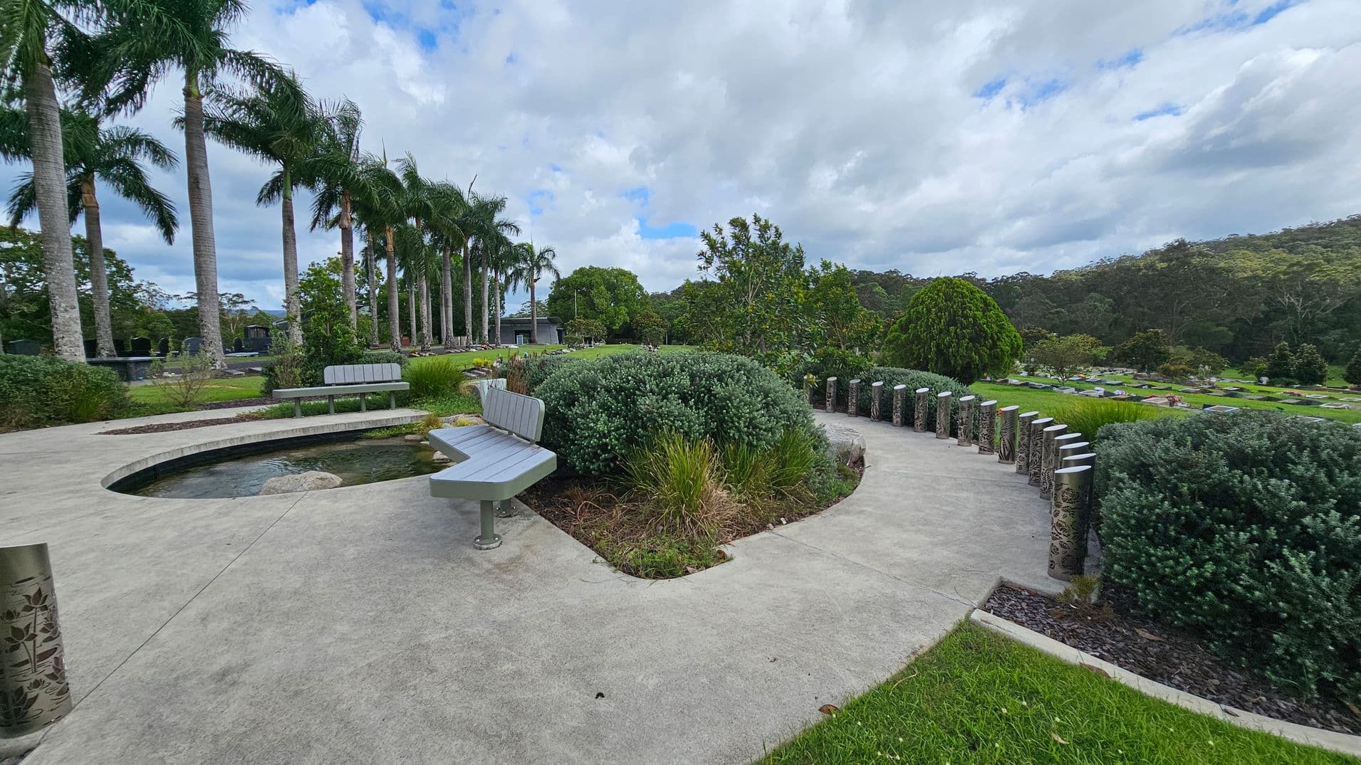 A landscaped garden with a curved concrete path and a small pond in the centre. On the left, there are tall palm trees lined up, and a few benches are placed along the path. Bushes and various plants are scattered throughout the area. On the right, decorative metal posts line the path, and more greenery extends into the distance. The background features a wooded area with dense trees.
