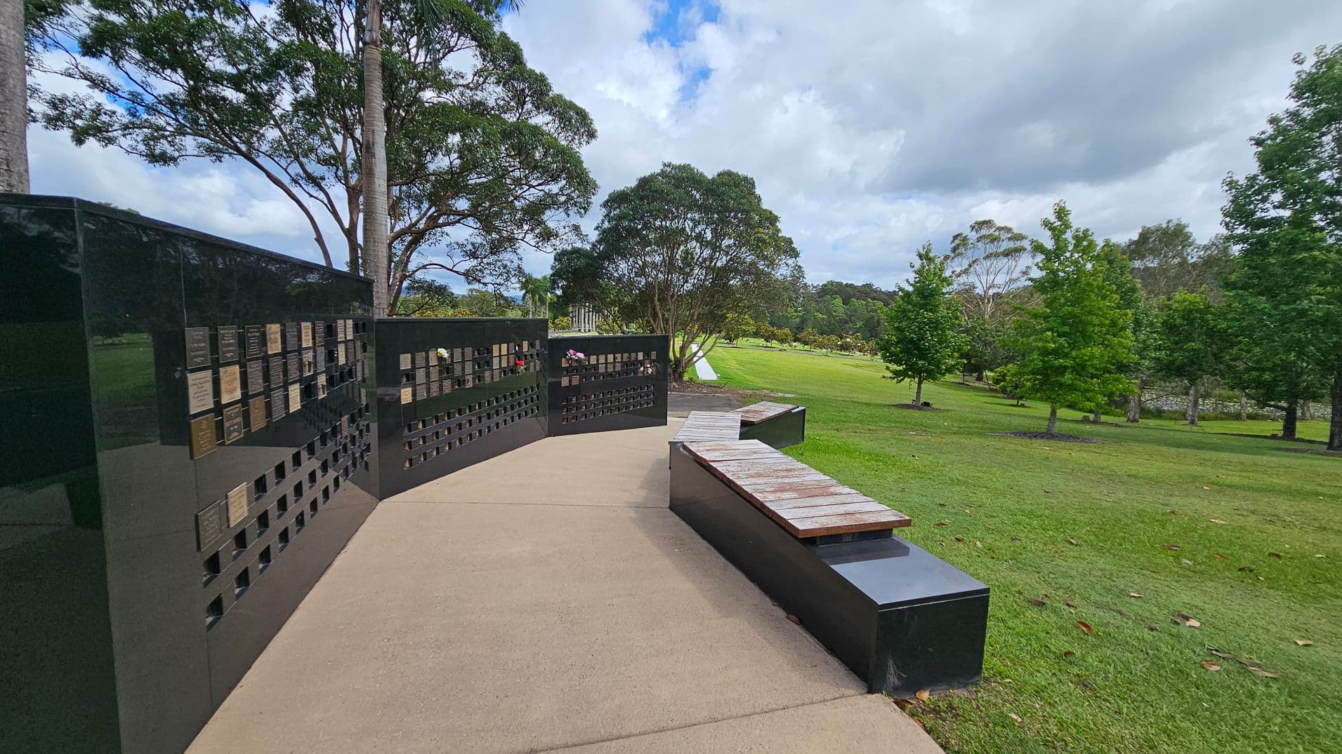 A paved pathway leads to a black memorial wall on the left, featuring numerous plaques. In front of the wall, there are wooden benches on black bases. The right side of the image shows a grassy area with scattered trees, including a large tree in the centre. The background is filled with more trees, creating a lush, green landscape.