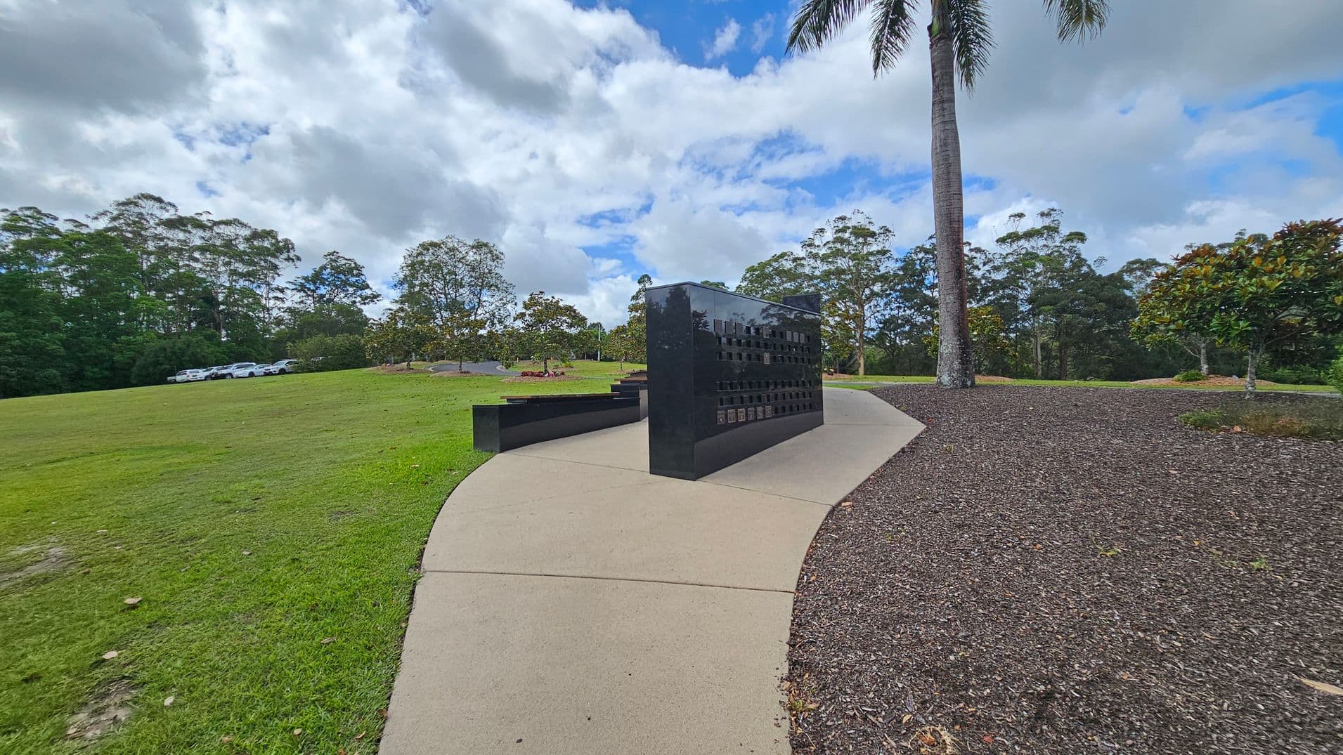 A black, polished memorial wall stands on a curved concrete path. To the left, there's a grassy area with several parked white cars in the distance. On the right, a tall palm tree and a small bush are situated on a bed of dark mulch. In the background, a line of trees borders the scene under a cloudy sky.