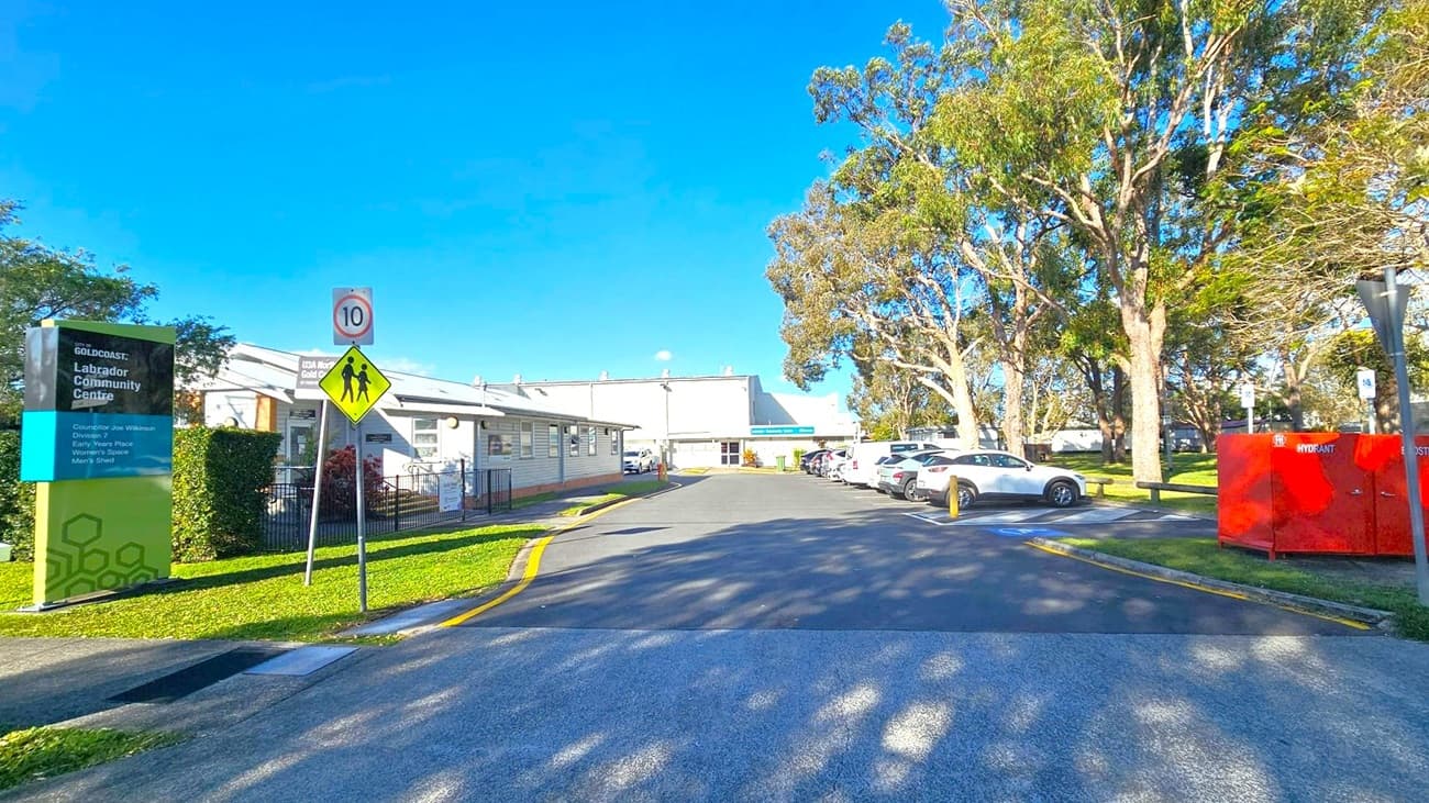 A paved road leads to a building in the centre, with a sign on the left reading "Labrador Community Centre." A speed limit sign and pedestrian crossing sign are nearby. On the right, several cars are parked under tall trees, and a red hydrant box is visible. The building on the left is single-storey with a light exterior. The area is surrounded by greenery, and the sky is clear and blue.